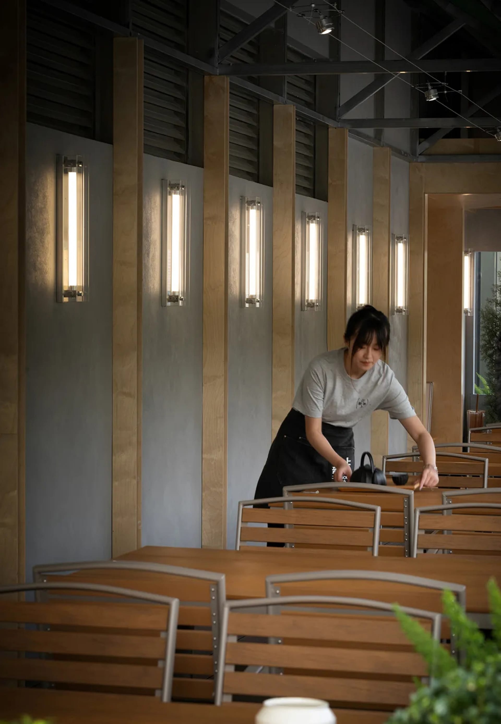 A woman cleaning a wooden table in a modern restaurant interior with geometric lighting fixtures.