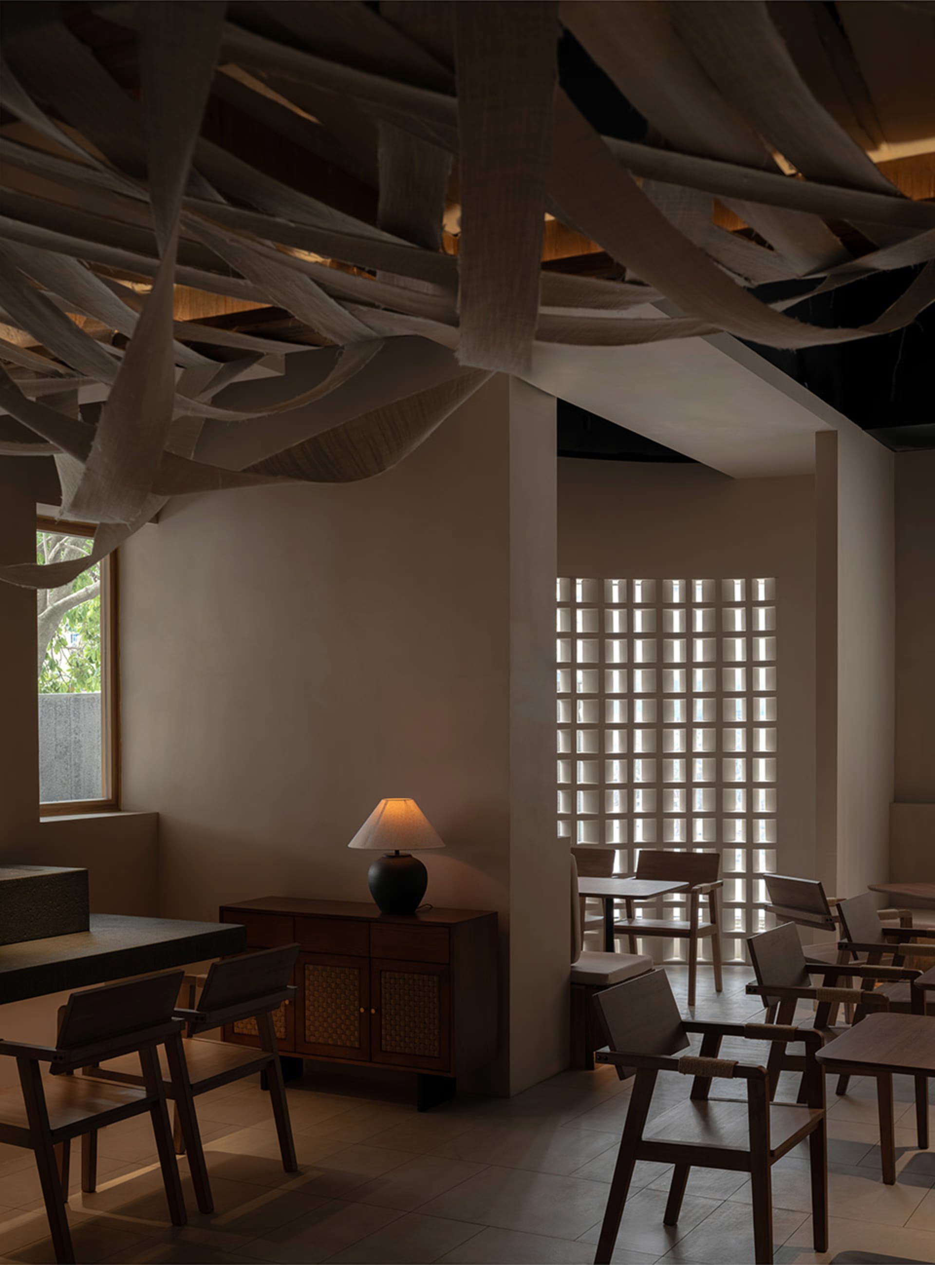 A minimalist restaurant interior featuring wooden tables, chairs, and a wooden cabinet with a lamp, with fabric-covered beams