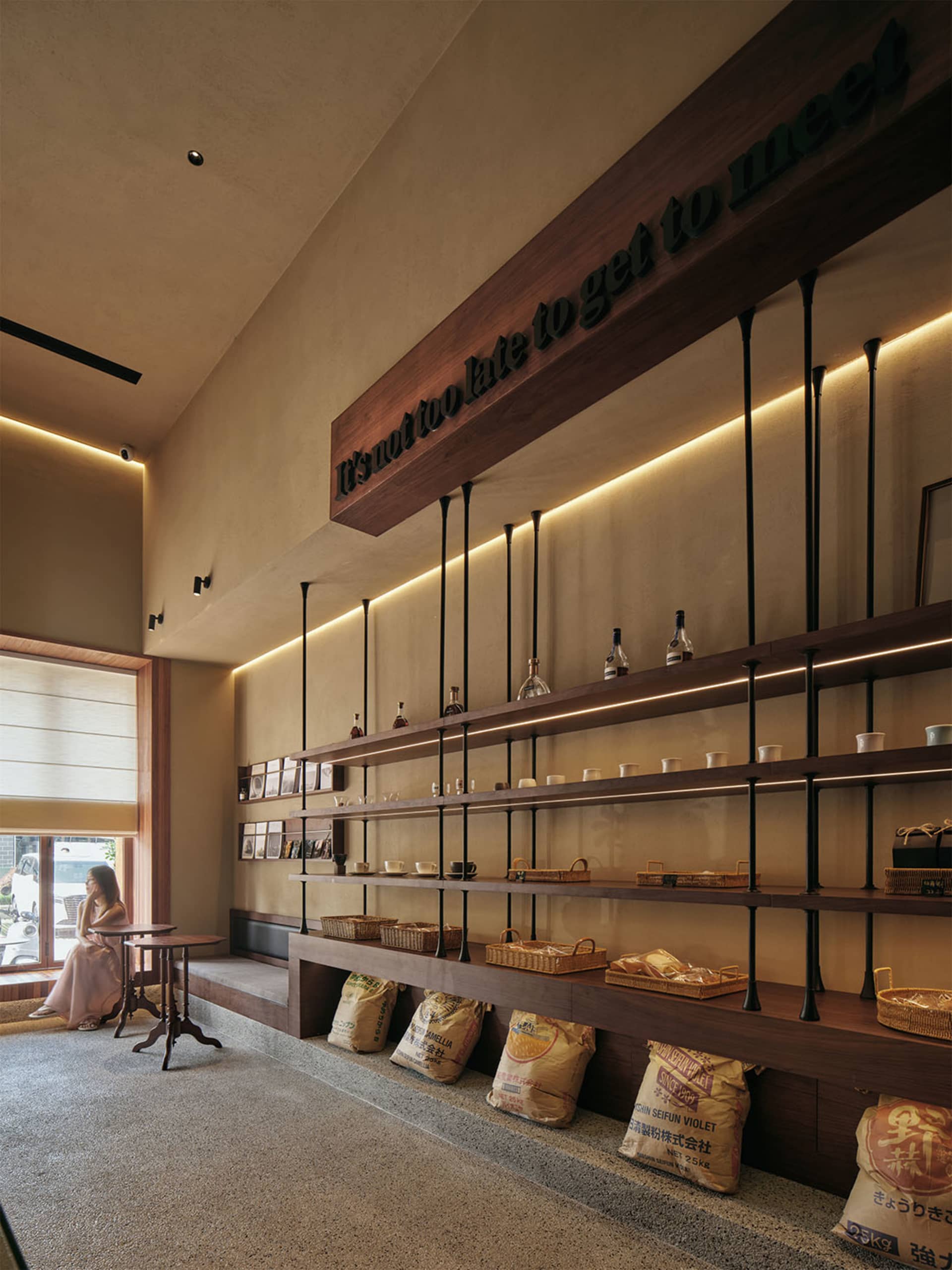 A cozy coffee shop with concrete shelves displaying various items and a woman sitting at a small table.