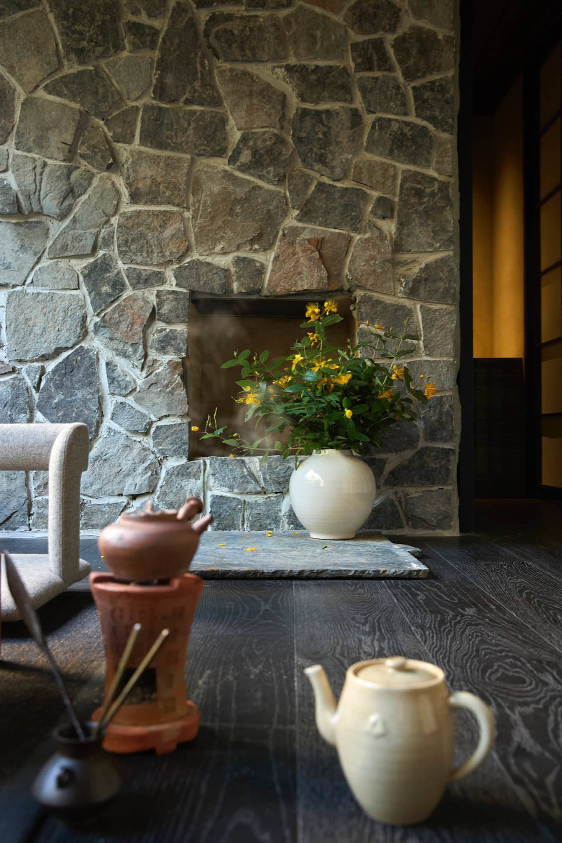 A cozy living room with a stone fireplace, wooden floor, and a white vase with yellow flowers.