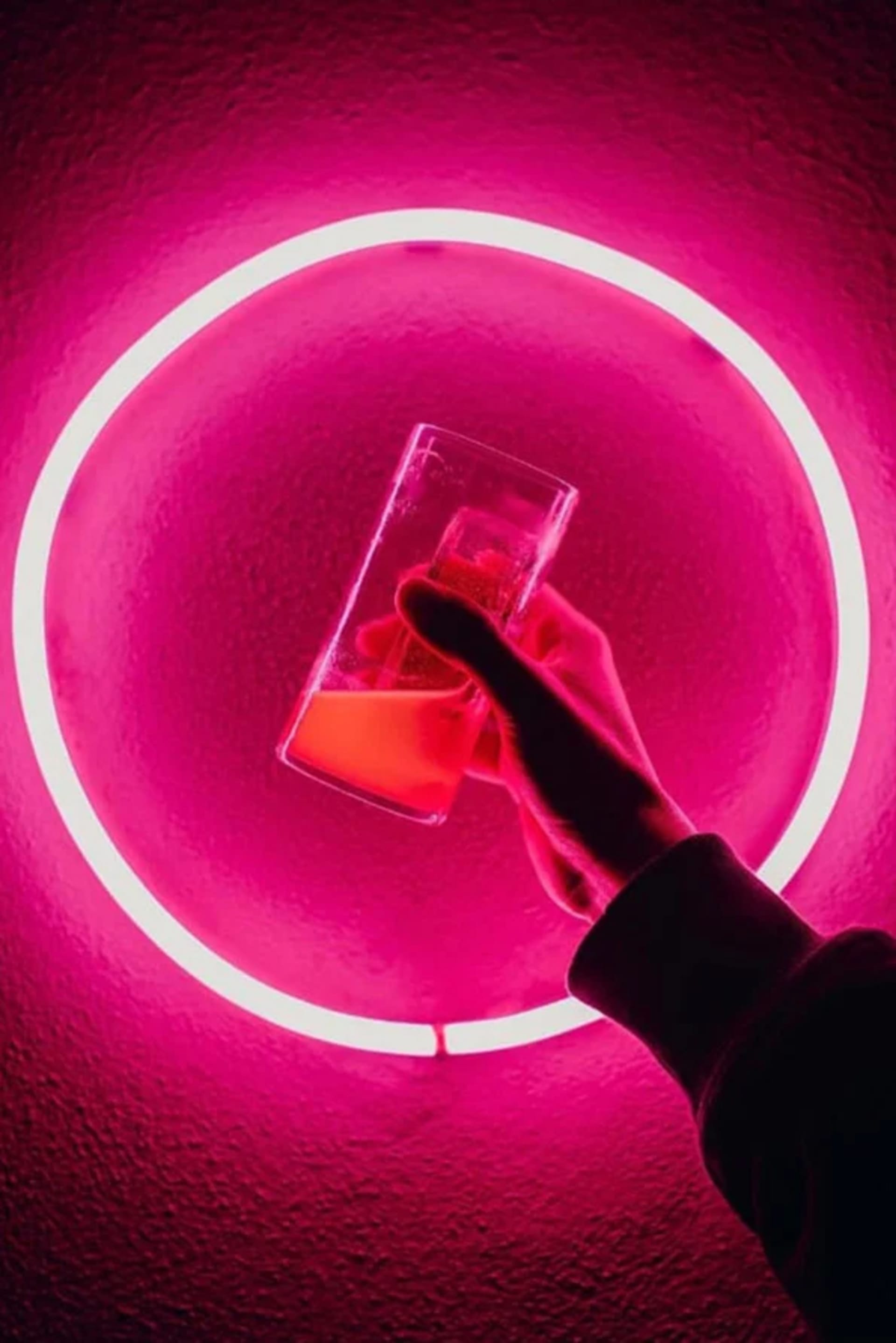 手握裝有粉紅液體的玻璃杯，背景為鮮豔的粉紅霓虹圓形。