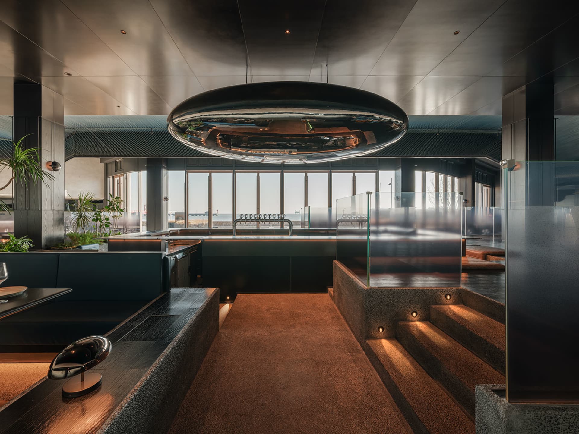 Modern restaurant interior with black metallic ceiling pendant, glass and concrete finishes, and minimalist design.