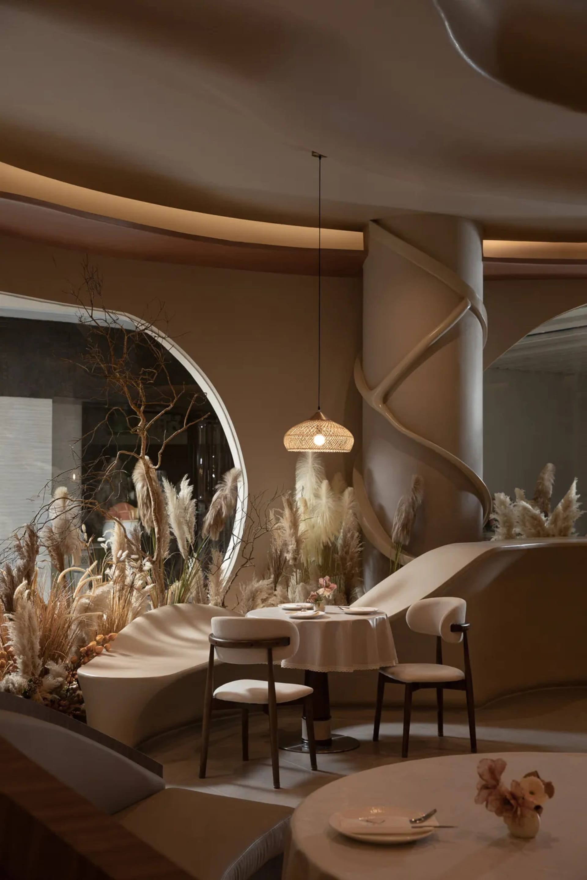 A modern restaurant interior with neutral textiles, featuring round tables, chairs, and decorative plants.
