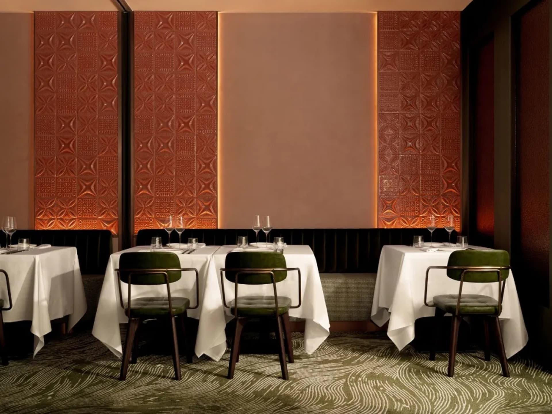A well-lit, modern restaurant dining room with textured brick walls, green velvet chairs, and white tablecloths.