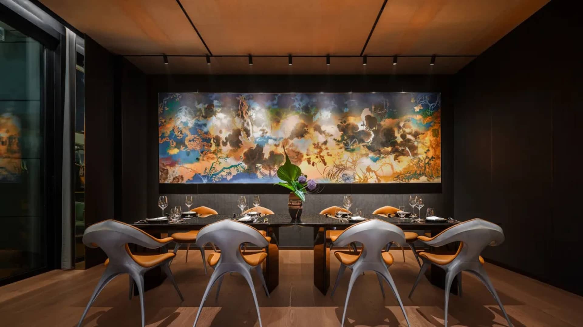 A sleek, industrial-style restaurant interior featuring metallic dining chairs, a wooden floor, and a large abstract mural.