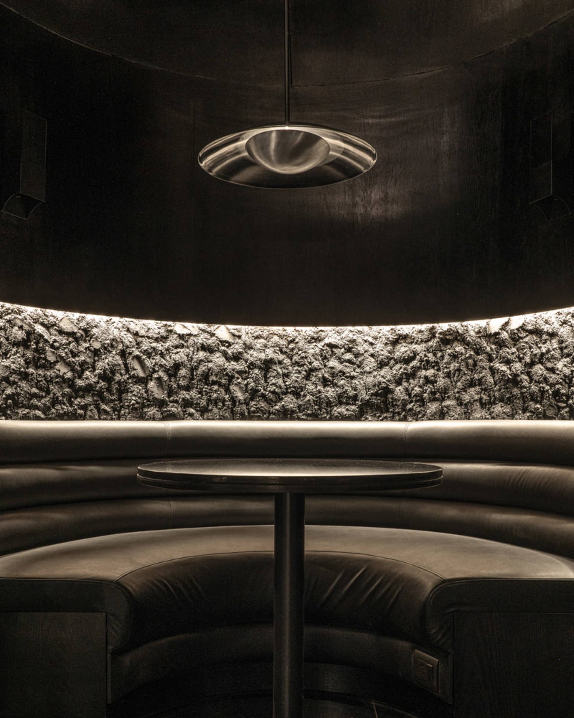 Circular booth with black cushions and a textured, illuminated concrete backdrop in a restaurant setting
