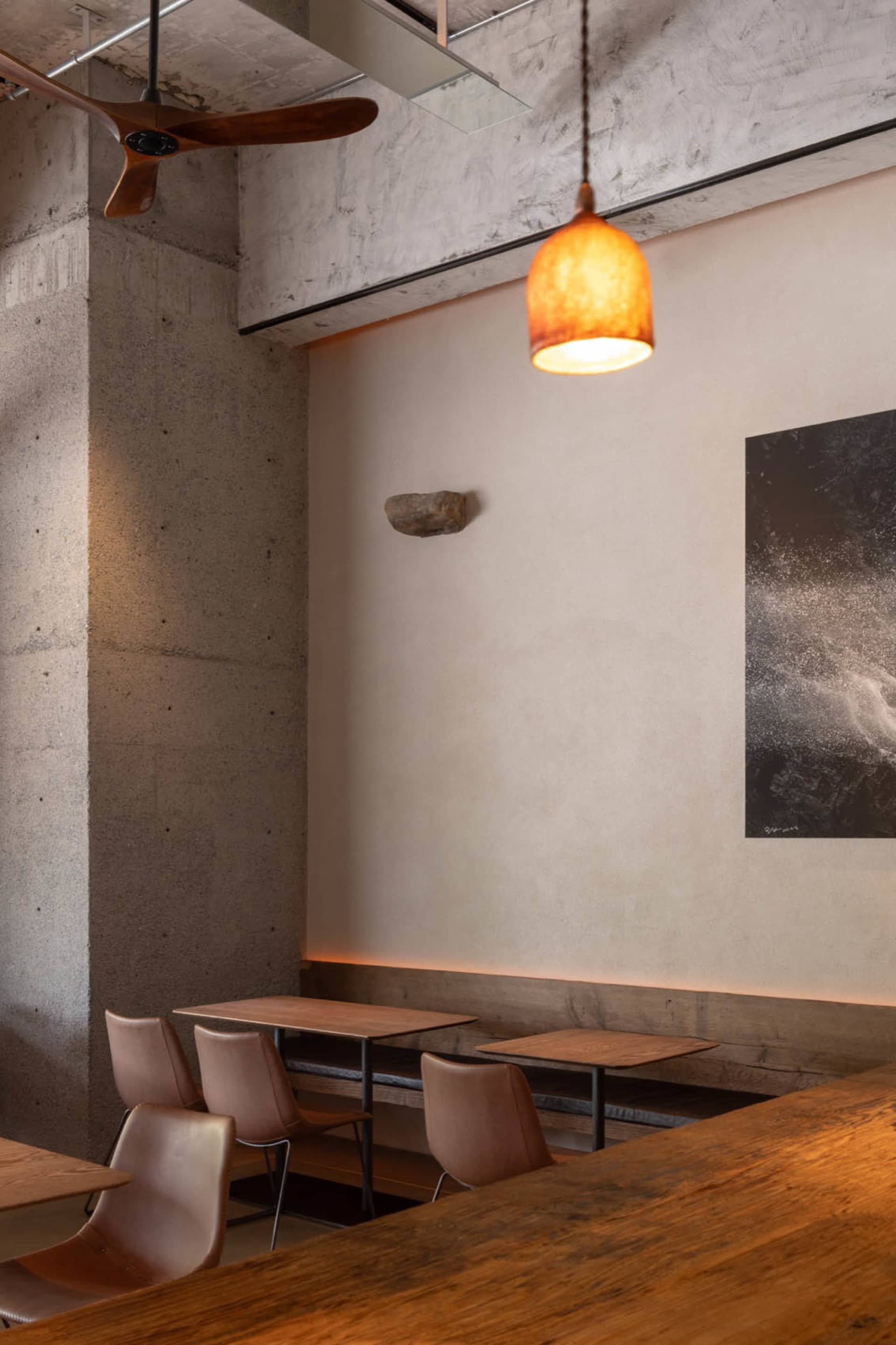 A modern, industrial-style restaurant with concrete and wooden elements, featuring a wooden ceiling fan and warm pendant ligh