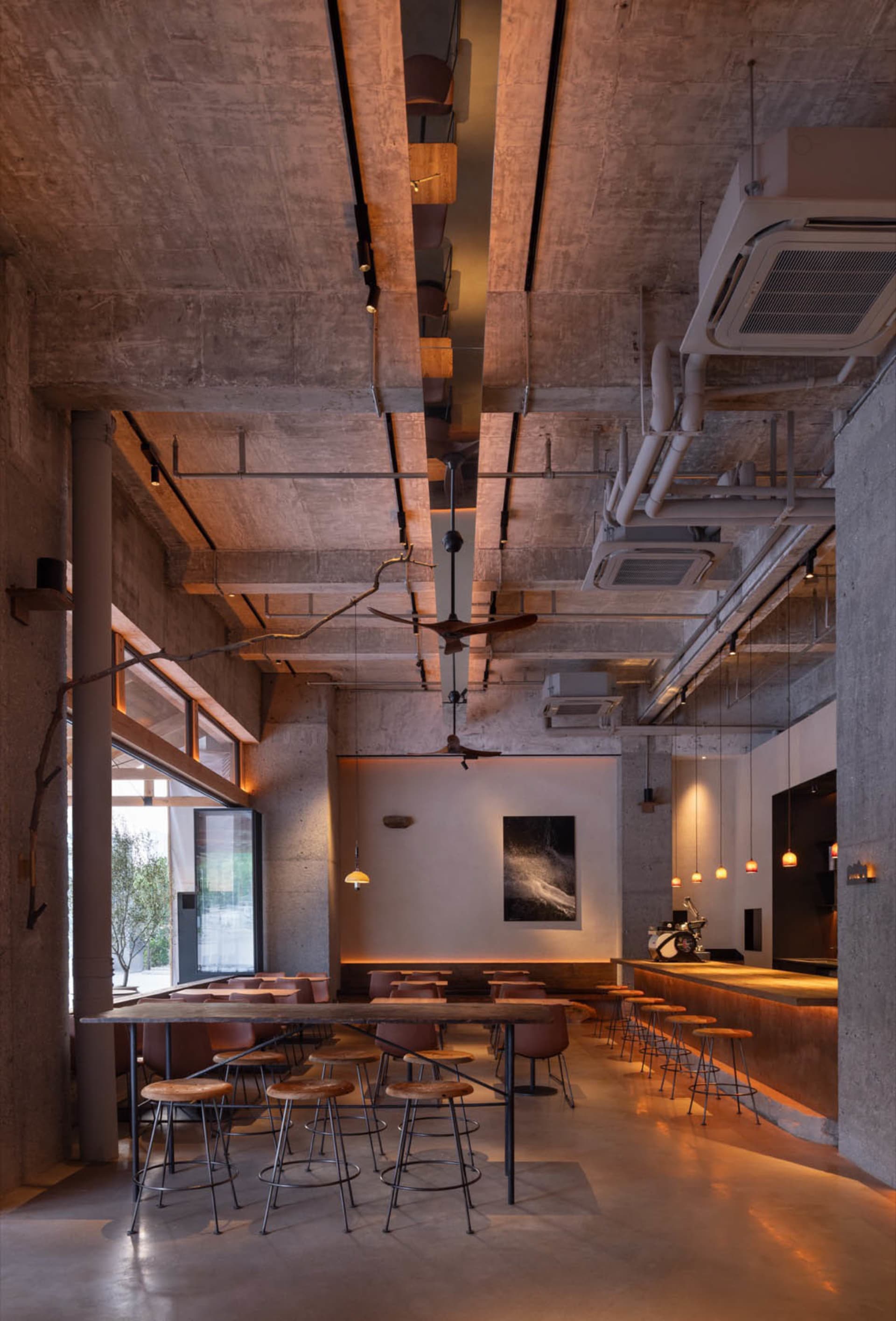 Industrial-style cafe interior with concrete walls, ceiling fans, and wooden stools.
