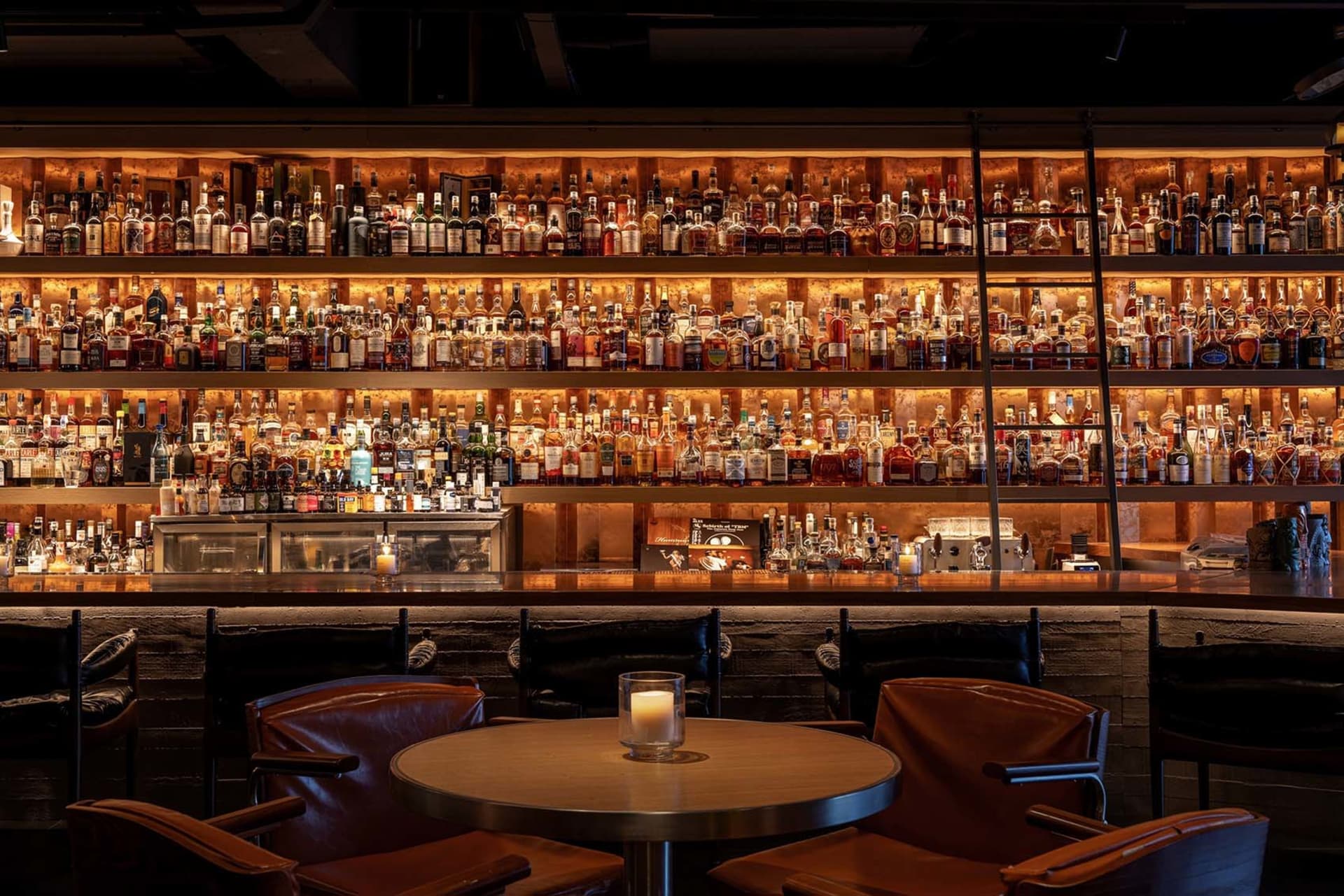 Industrial-style restaurant bar with wooden seating, metal tables, and glass bottles in the background.