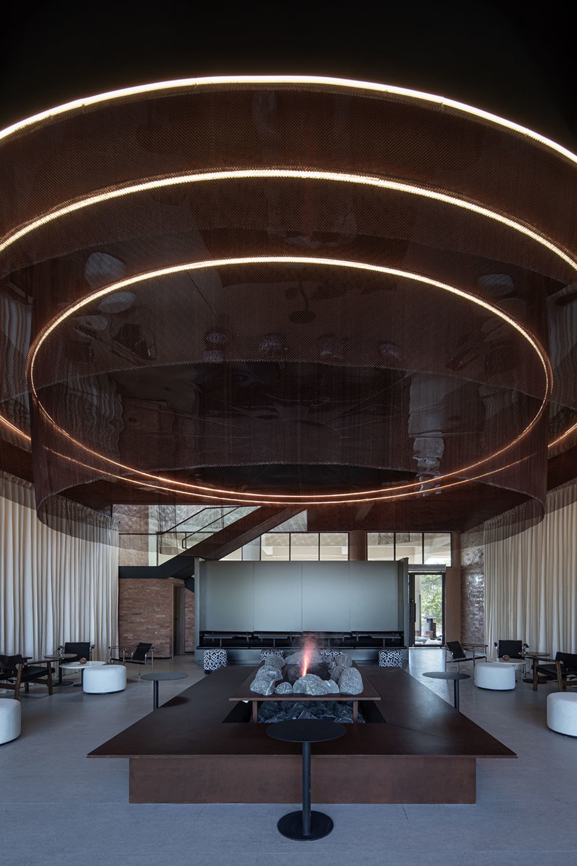 An industrial-style lobby with brick walls, metal railings, and a glass ceiling, featuring a central fire pit.