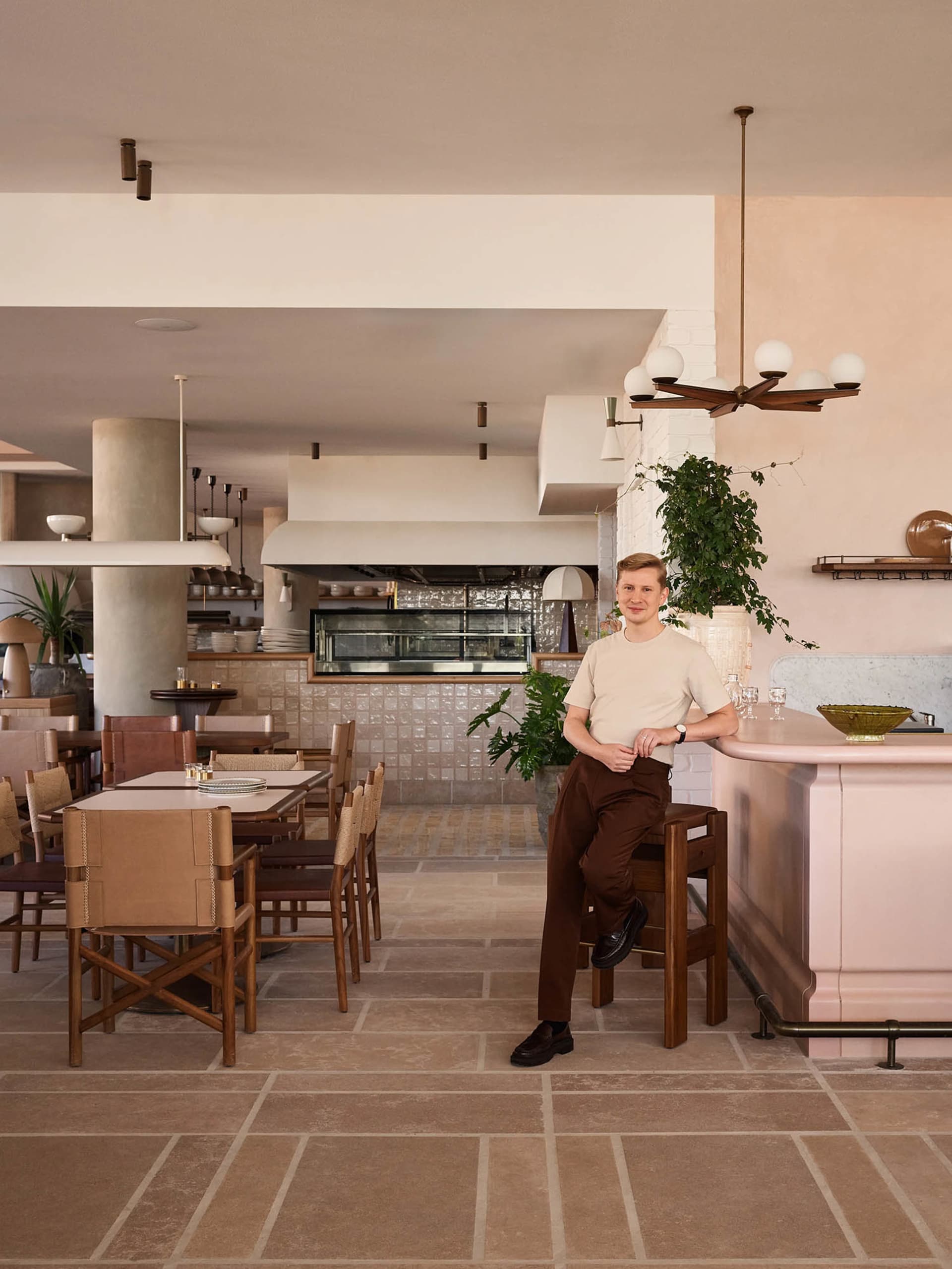 A man wearing a beige shirt and brown pants is seated at the bar of a modern restaurant, with clean lines and a minimalist ae