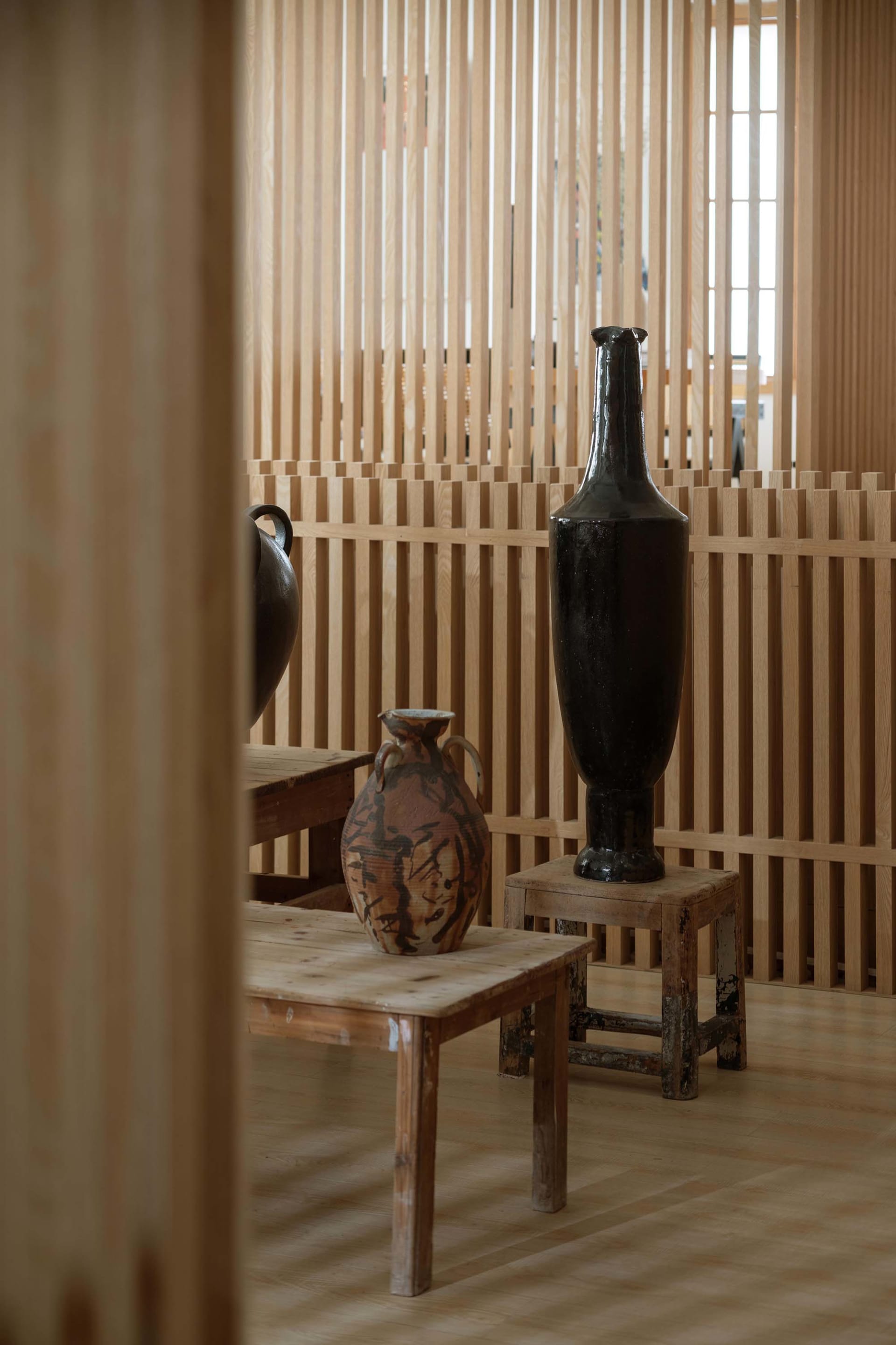 Decorative ceramic vases placed on wooden stools and tables in a room with wooden slat walls.