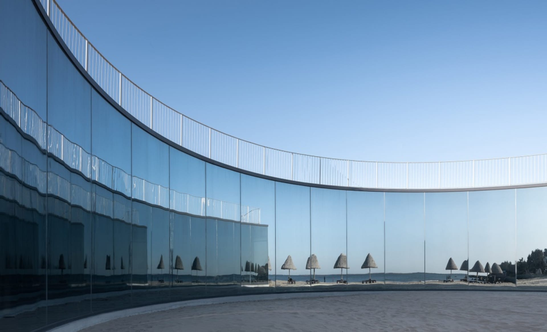 A modern, curved glass installation reflecting a beach scene and beach huts.