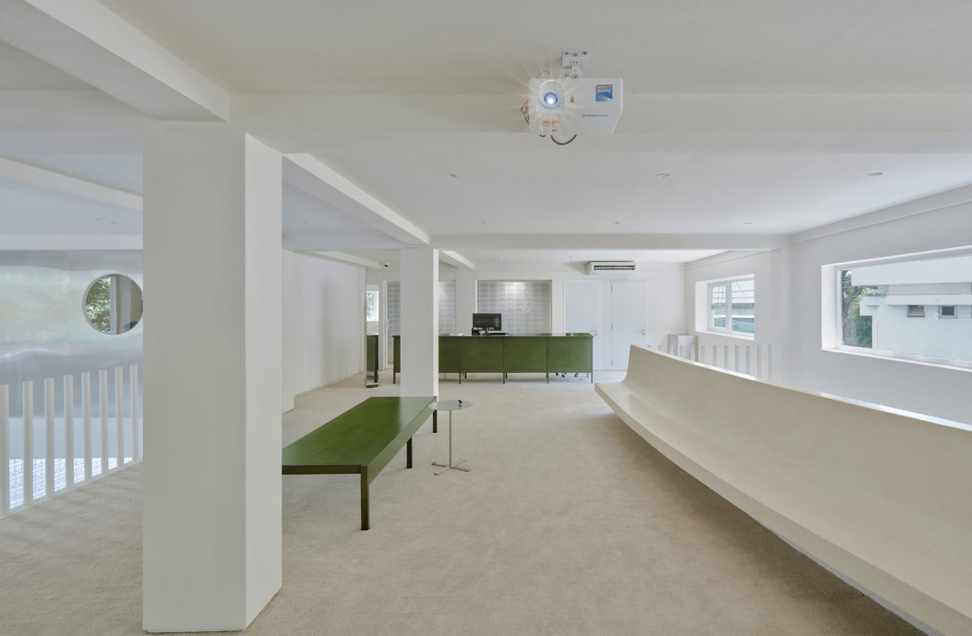 A modern office space featuring white concrete walls, a green reception desk, and minimalist furniture.