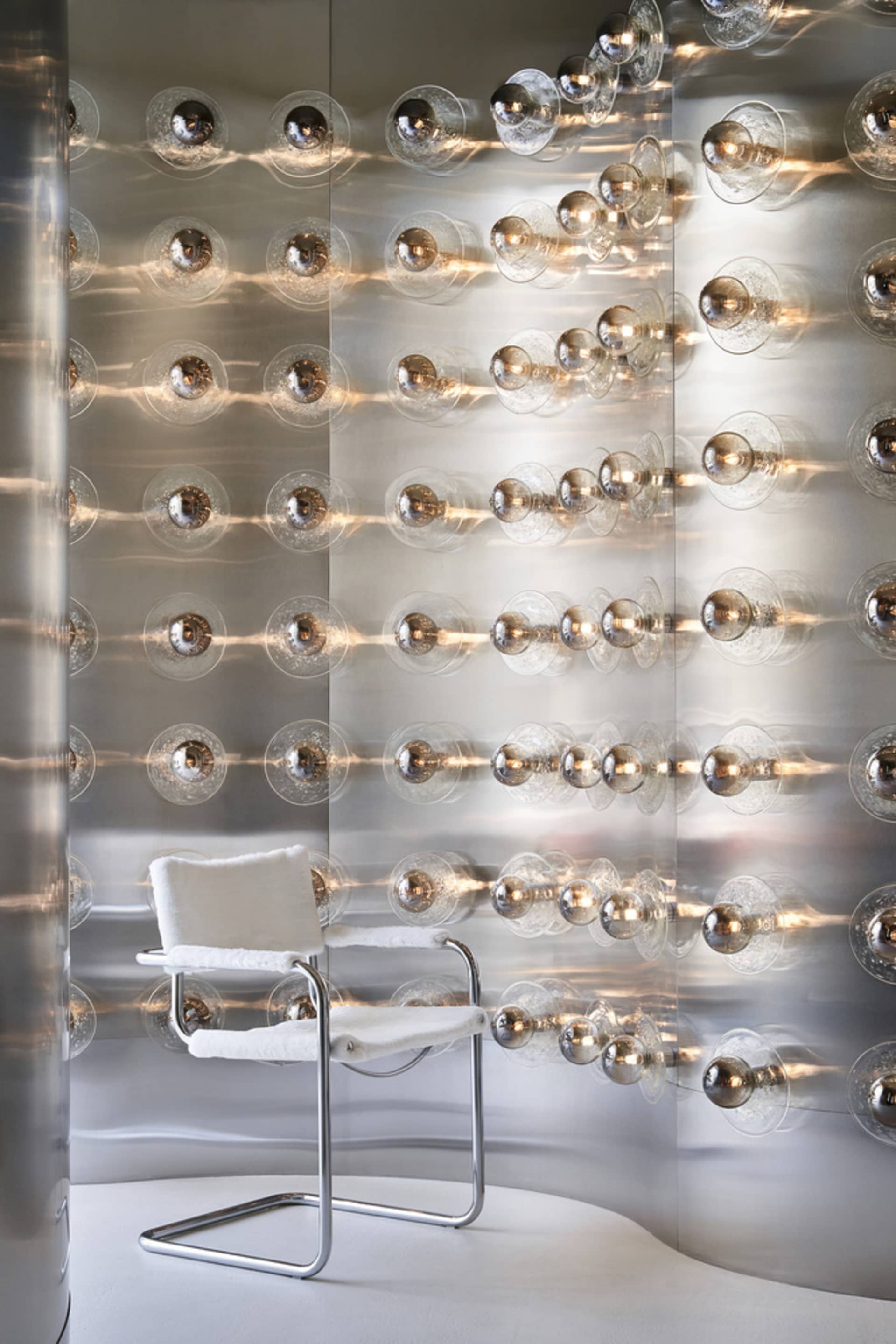 A minimalist, modern interior featuring a metallic wall with circular elements and a white chair with a cushion.