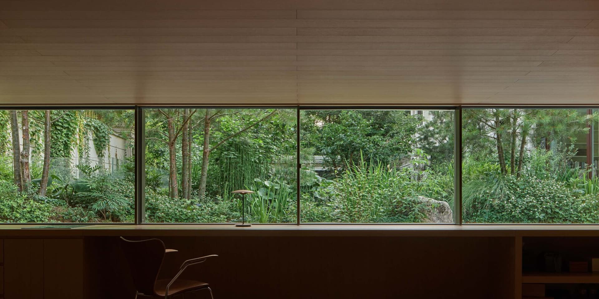 A minimalist office space featuring wooden desks and shelves, with large windows and greenery outside.