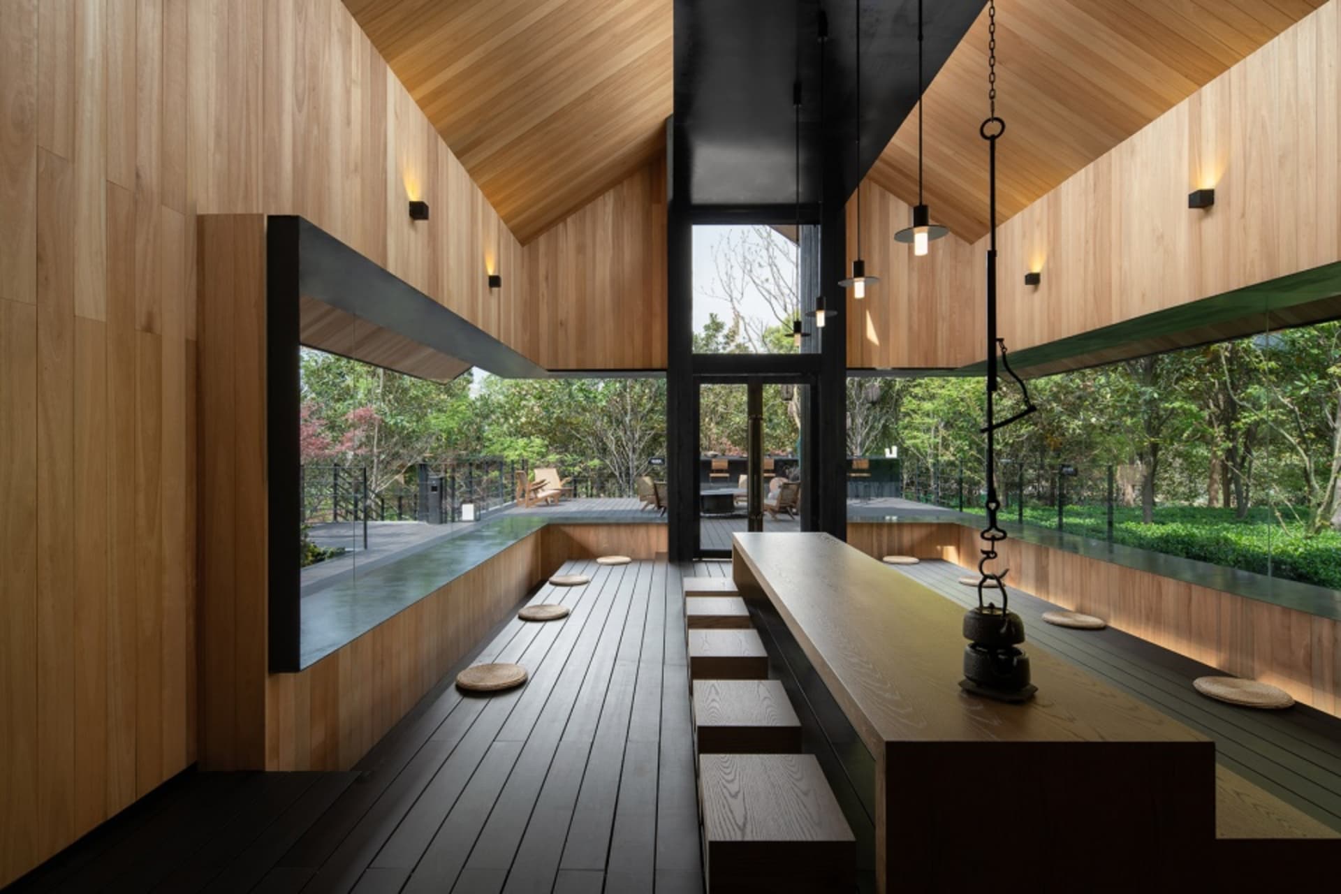 A minimalist restaurant interior with wooden tables and benches, surrounded by large windows overlooking greenery.
