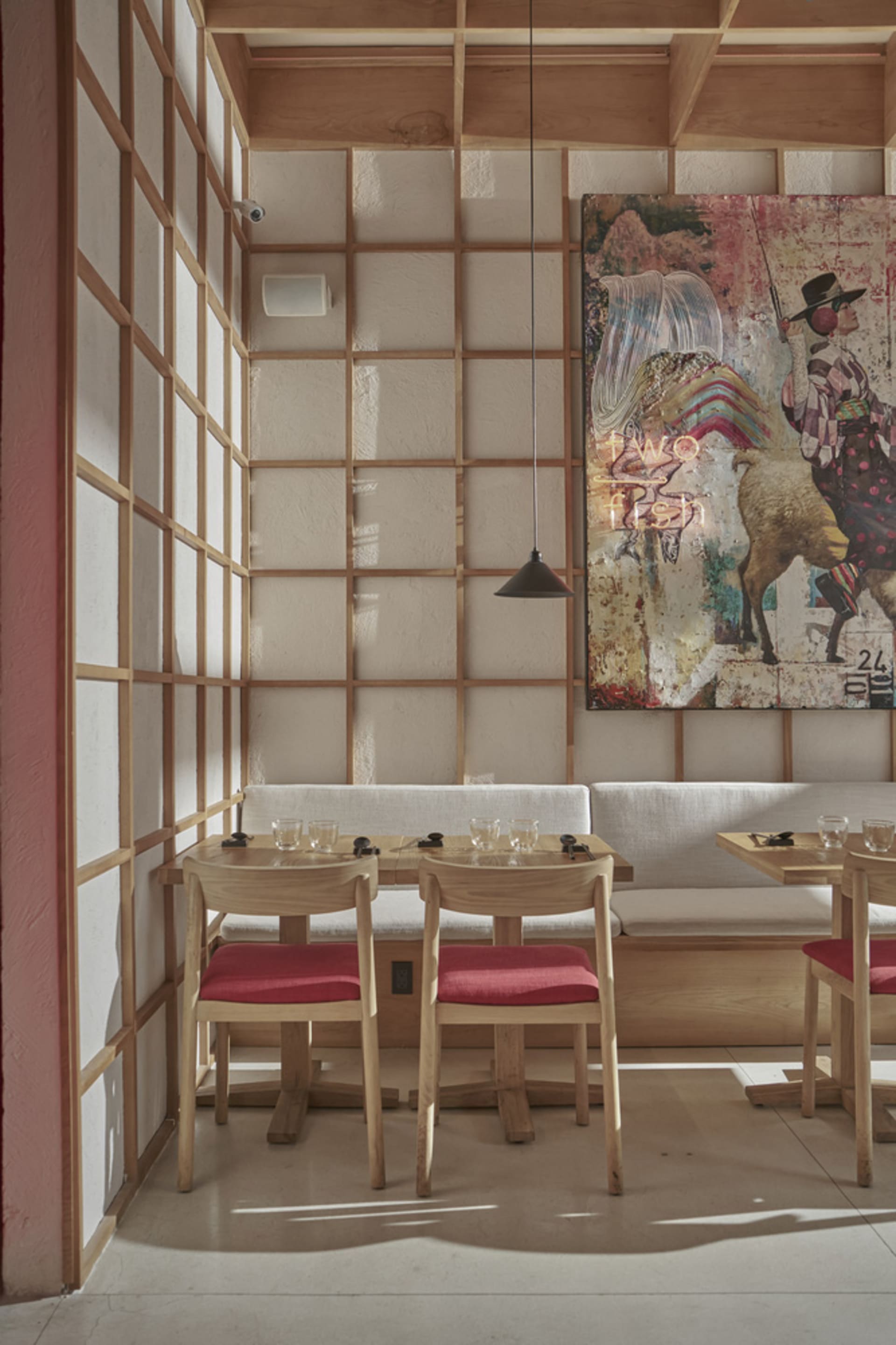 Modern restaurant interior with wooden tables and red cushions, featuring a colorful artwork on the wall.