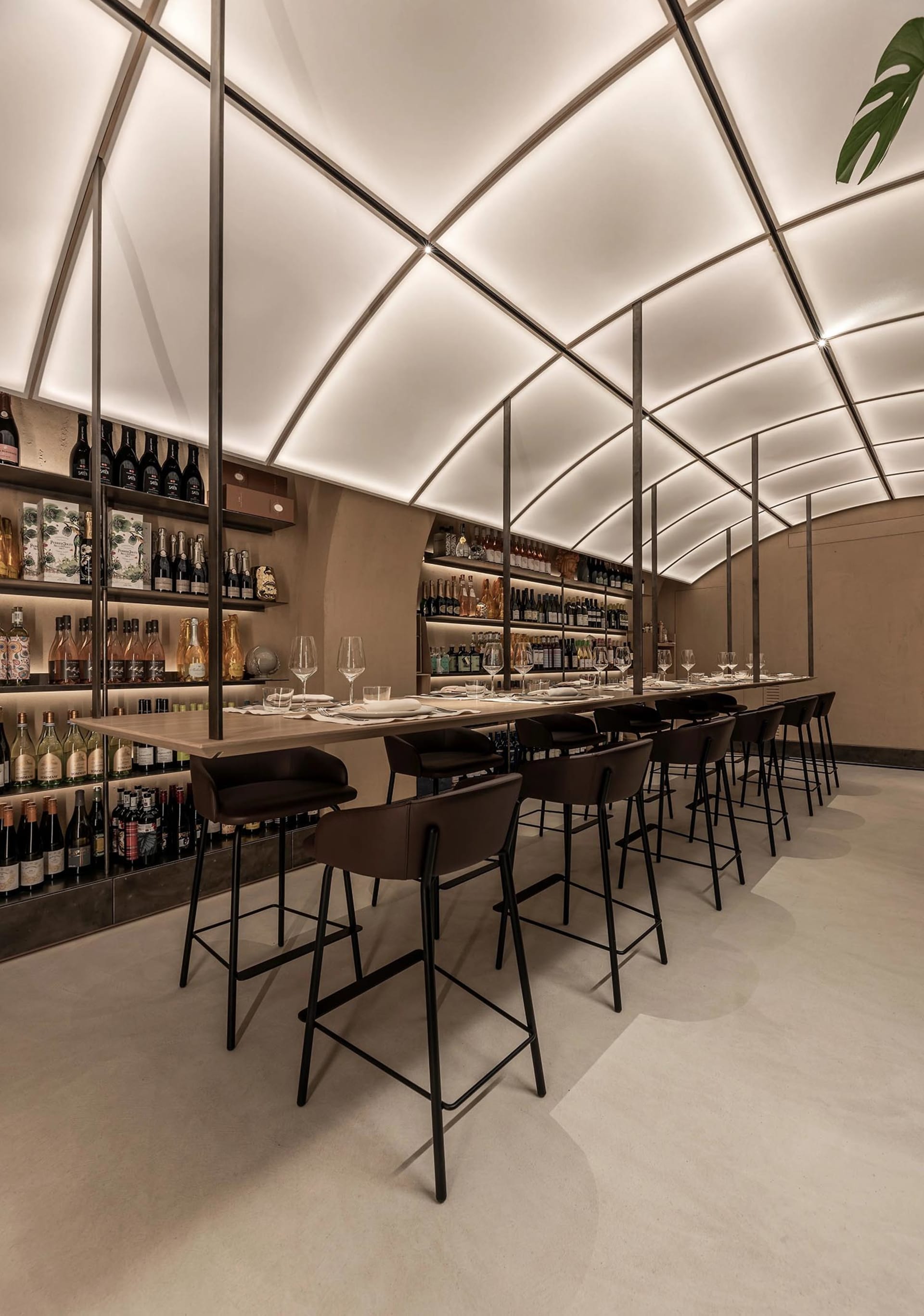 A modern restaurant bar with sleek design elements, including a long table with seating, wine bottles on shelves, and a conte