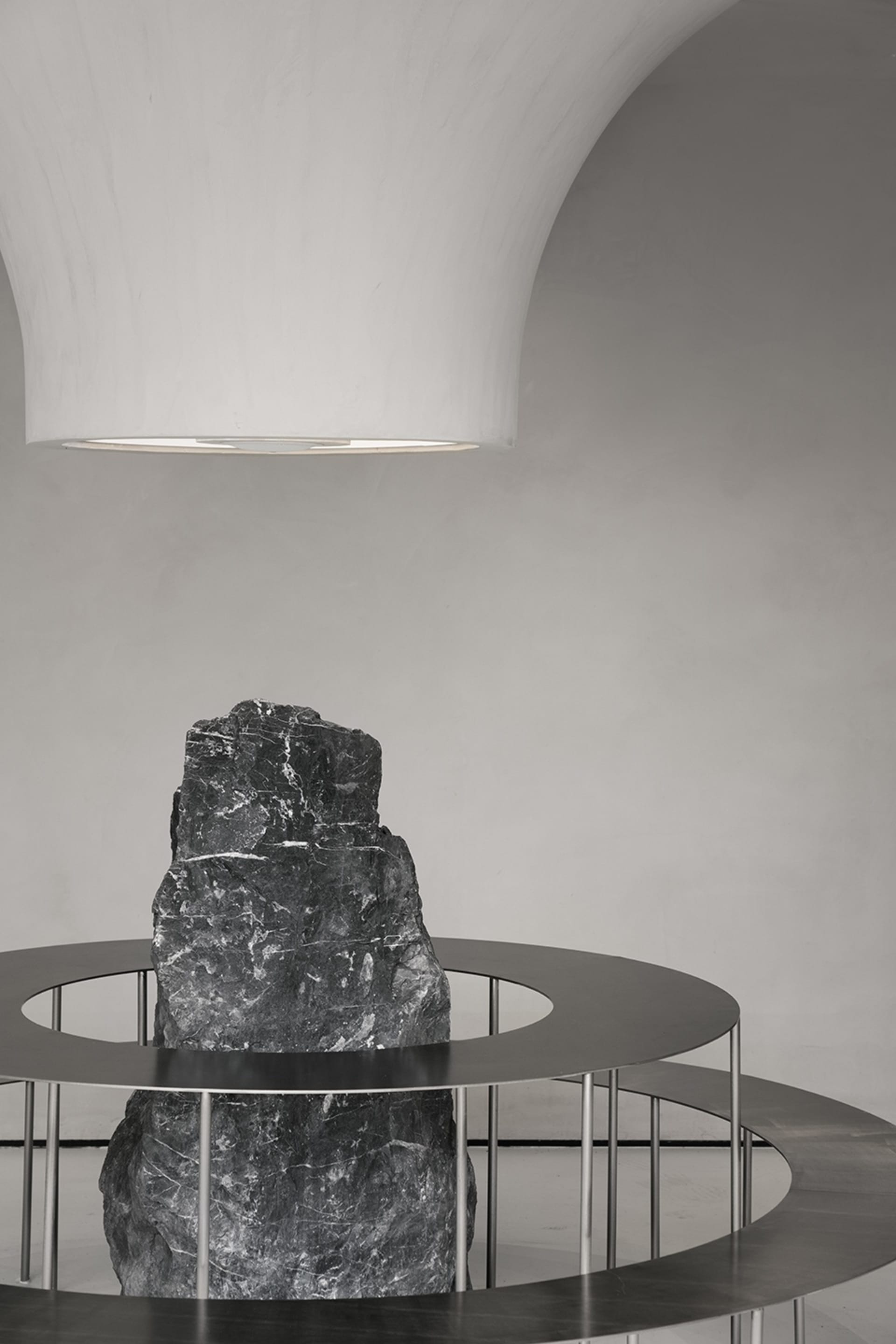 A black rock-like object encased in a circular metal frame, illuminated by a large, white ceiling light in a minimalist indus