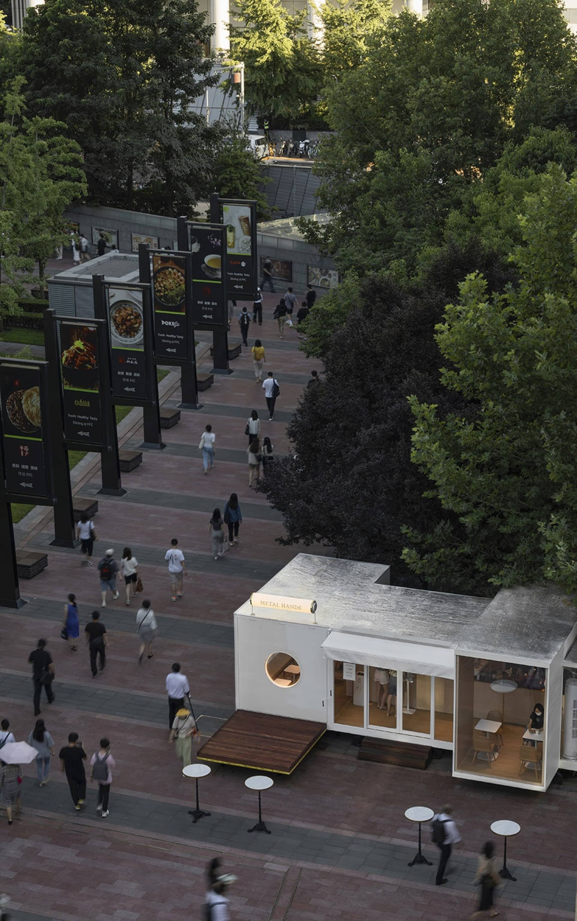 Outdoor area of a street-side cafe with metal exterior, surrounded by greenery and pedestrian walkways.