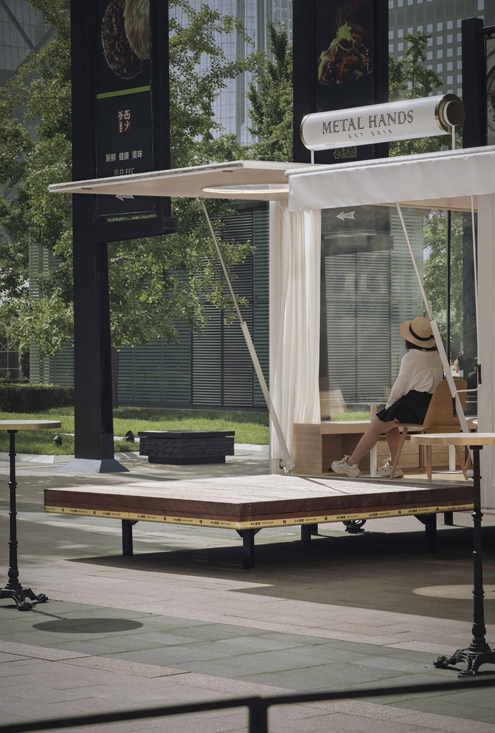 A minimalist urban cafe with wooden platforms, metal seating, glass walls, and fabric curtains.
