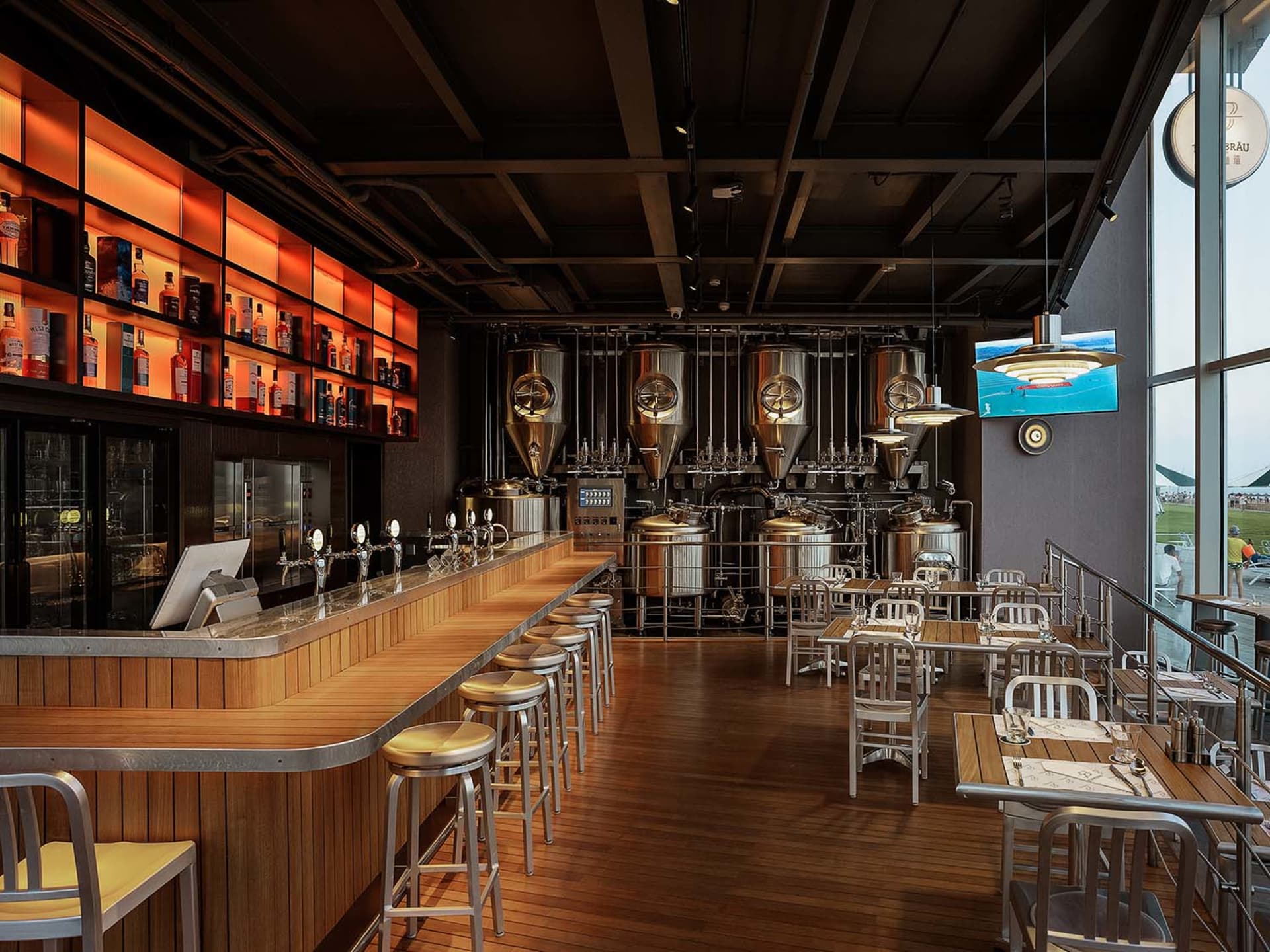 Industrial-themed restaurant bar with wooden tables, metal stools, and glass brewing tanks.