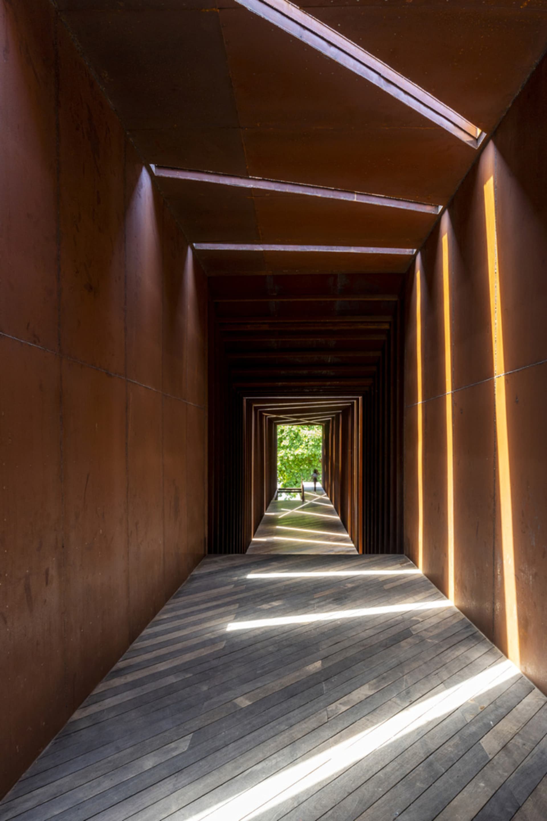 A narrow hallway lined with industrial-style wood flooring and metallic walls, leading to an open area with greenery.