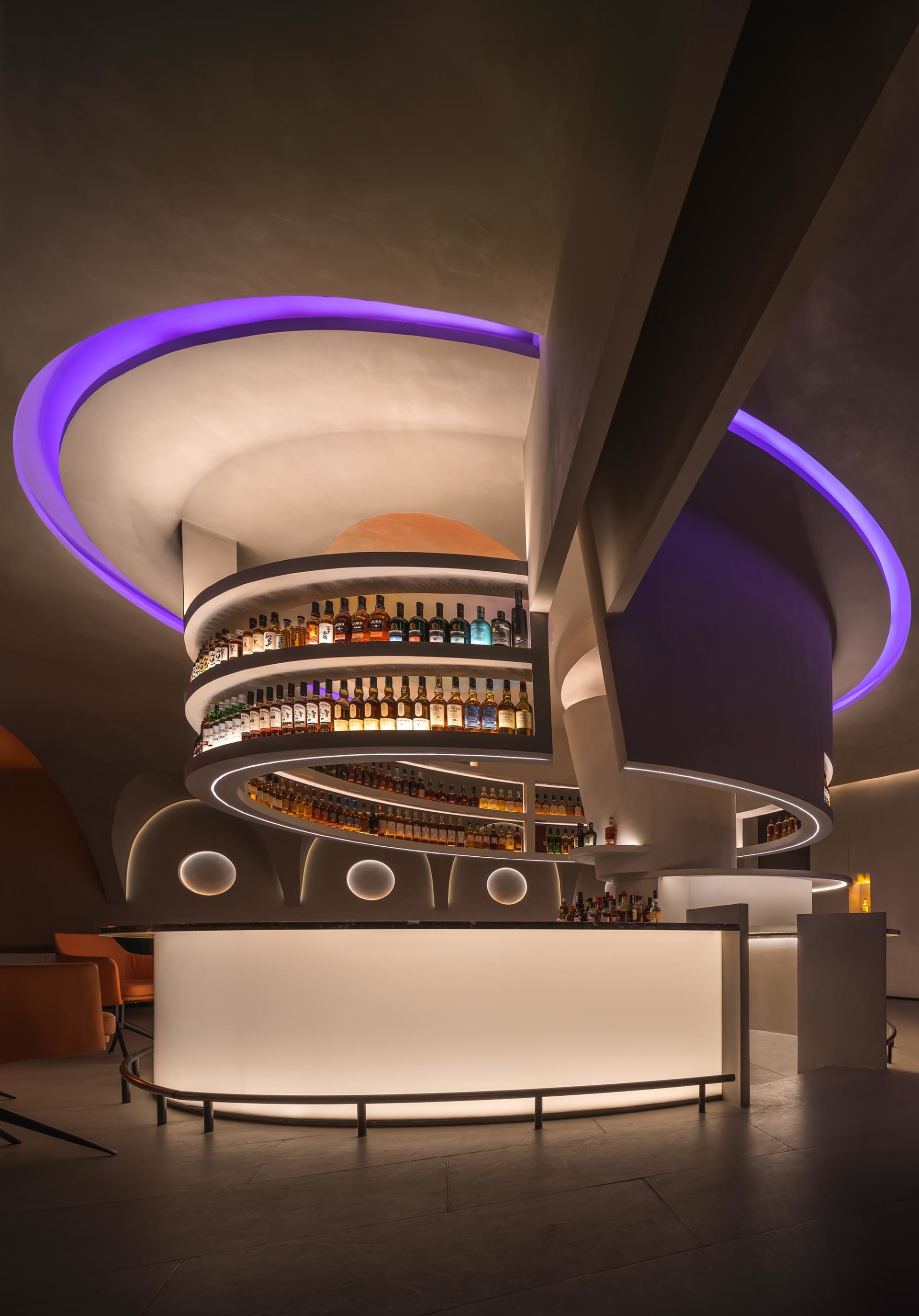 A modern bar with shelves stocked with bottles of alcohol, illuminated by soft lighting and accented with purple LED lights.