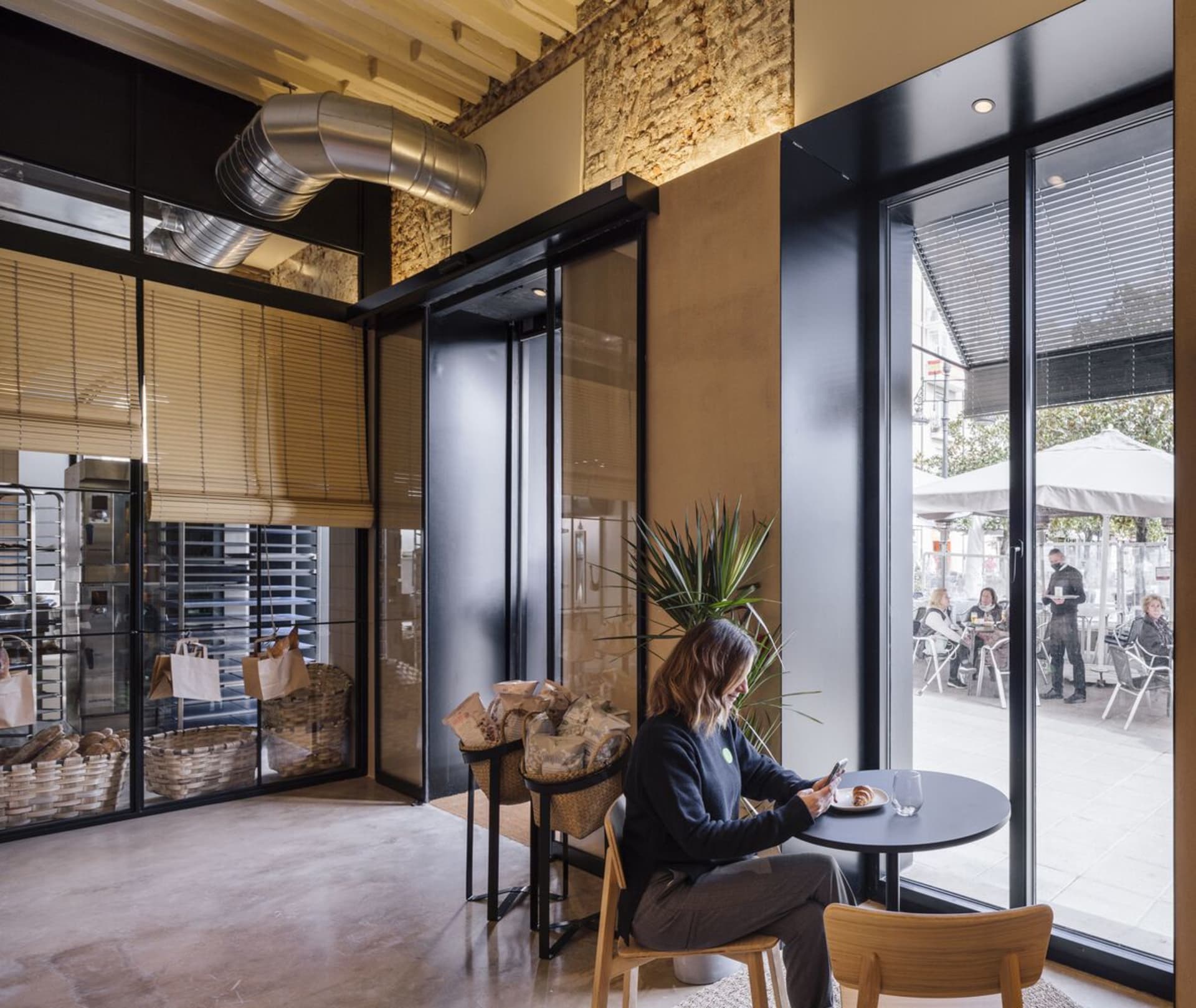 A person sitting at a small table in an industrial restaurant with metal pipes and glass walls.
