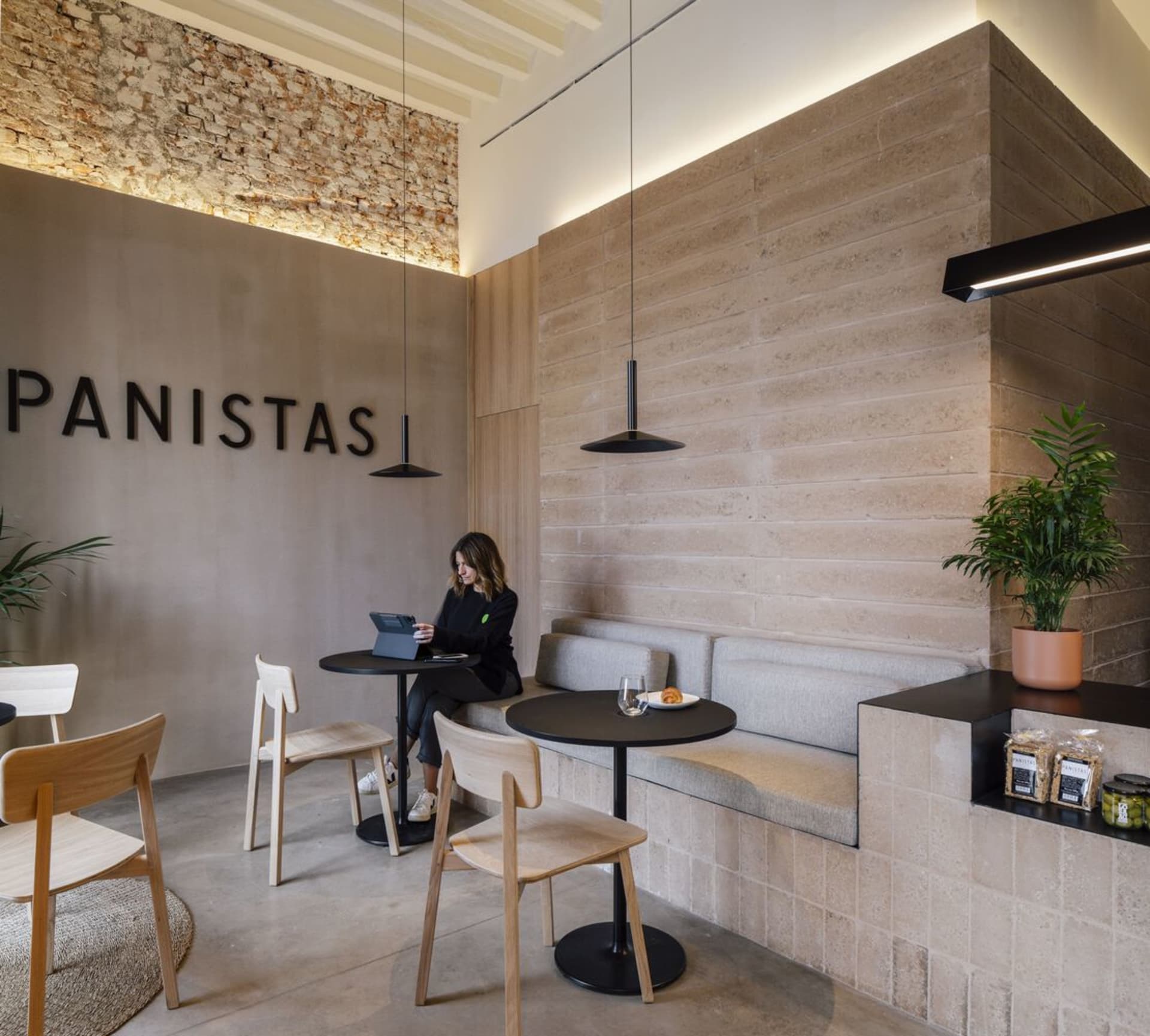A minimalist cafe with concrete and wood materials, featuring seating, tables, and a wall inscription that says "Panistas."