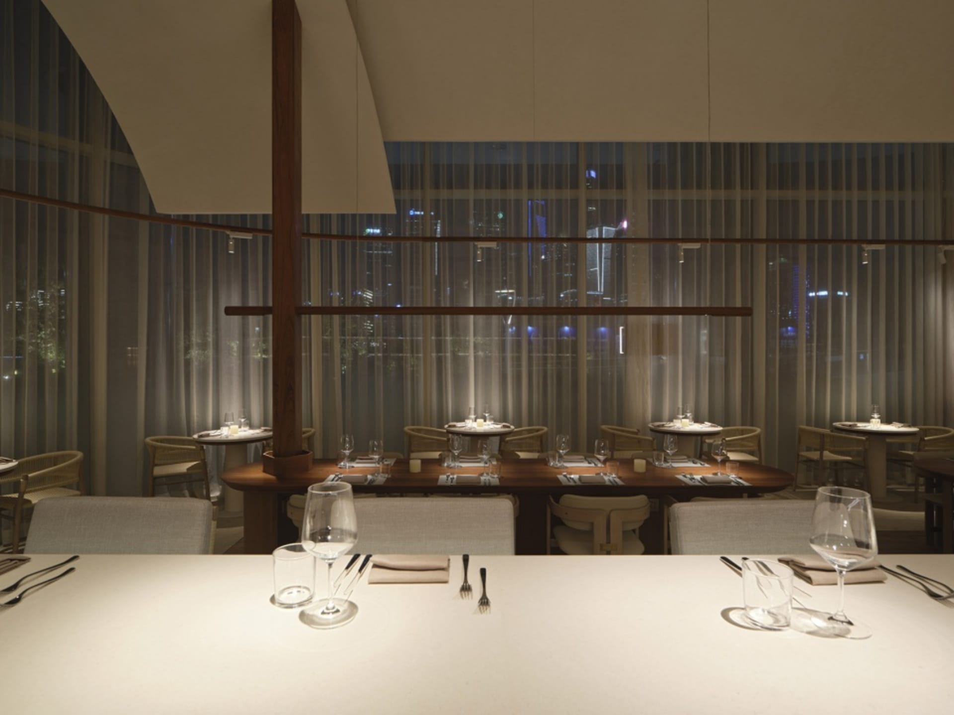 A modern dining room with glass and wooden furniture, set tables with white tablecloths and silverware, and large windows sho