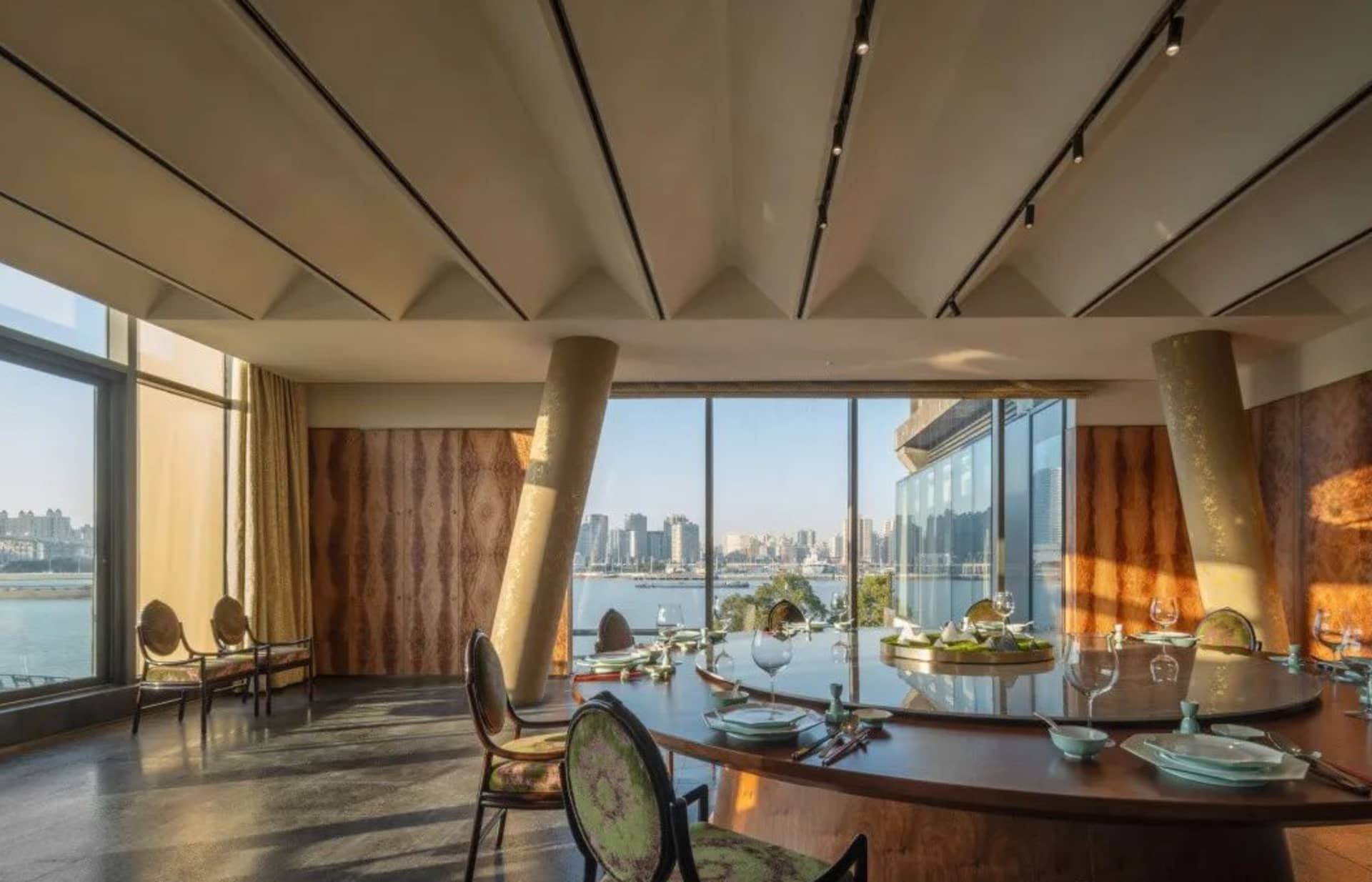A stylish restaurant interior featuring metallic columns, a large glass window overlooking a cityscape, and a dining table se