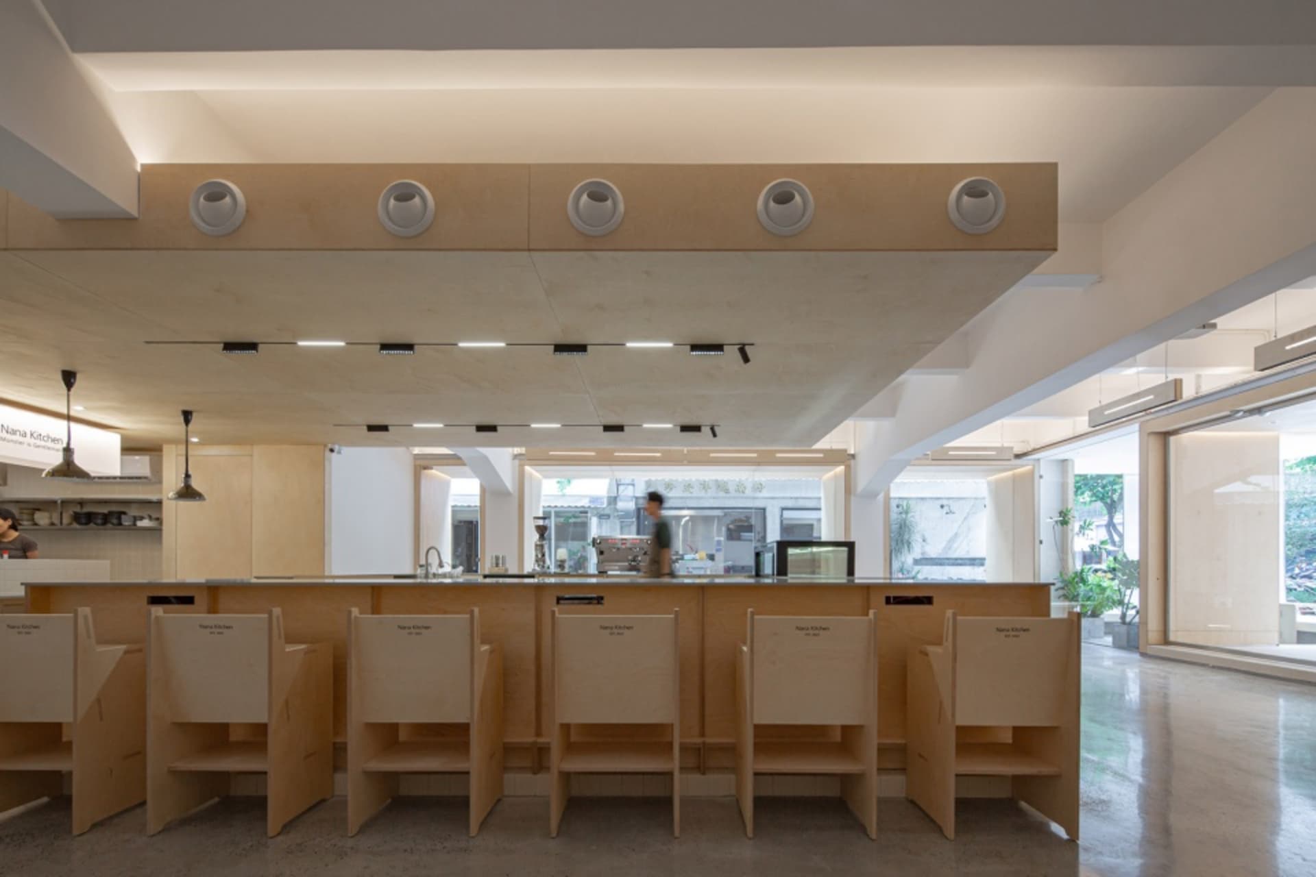 A minimalist restaurant/bar interior with wooden tables and chairs, open kitchen, and large windows.