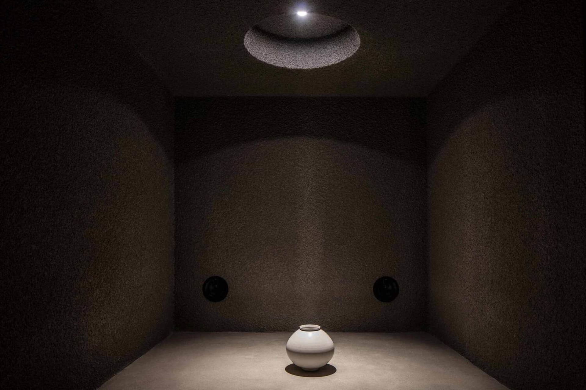 Minimalist interior space with a illuminated white ceramic vase on a wooden floor.