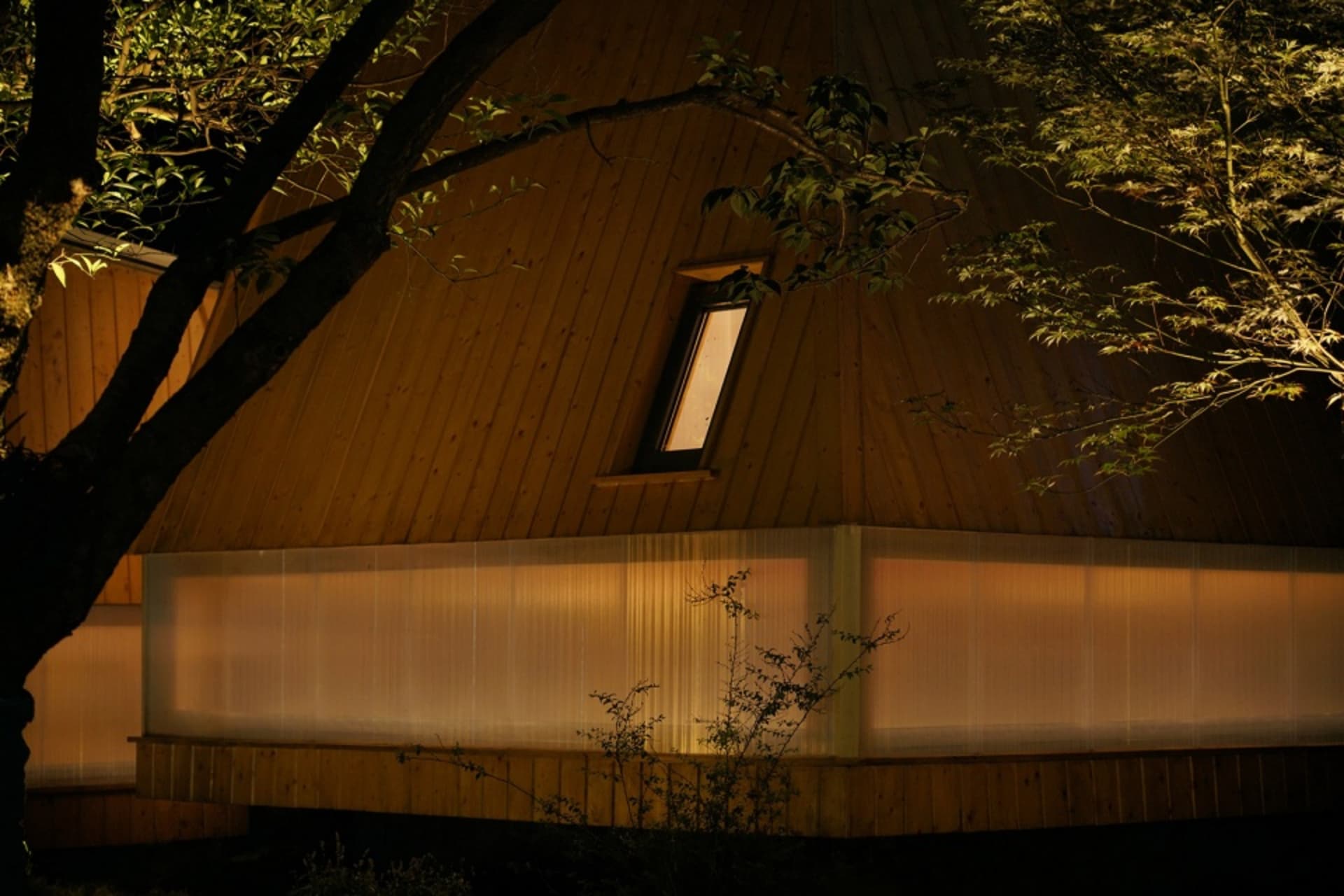 A modern house at night, featuring wooden exterior and a single illuminated window.