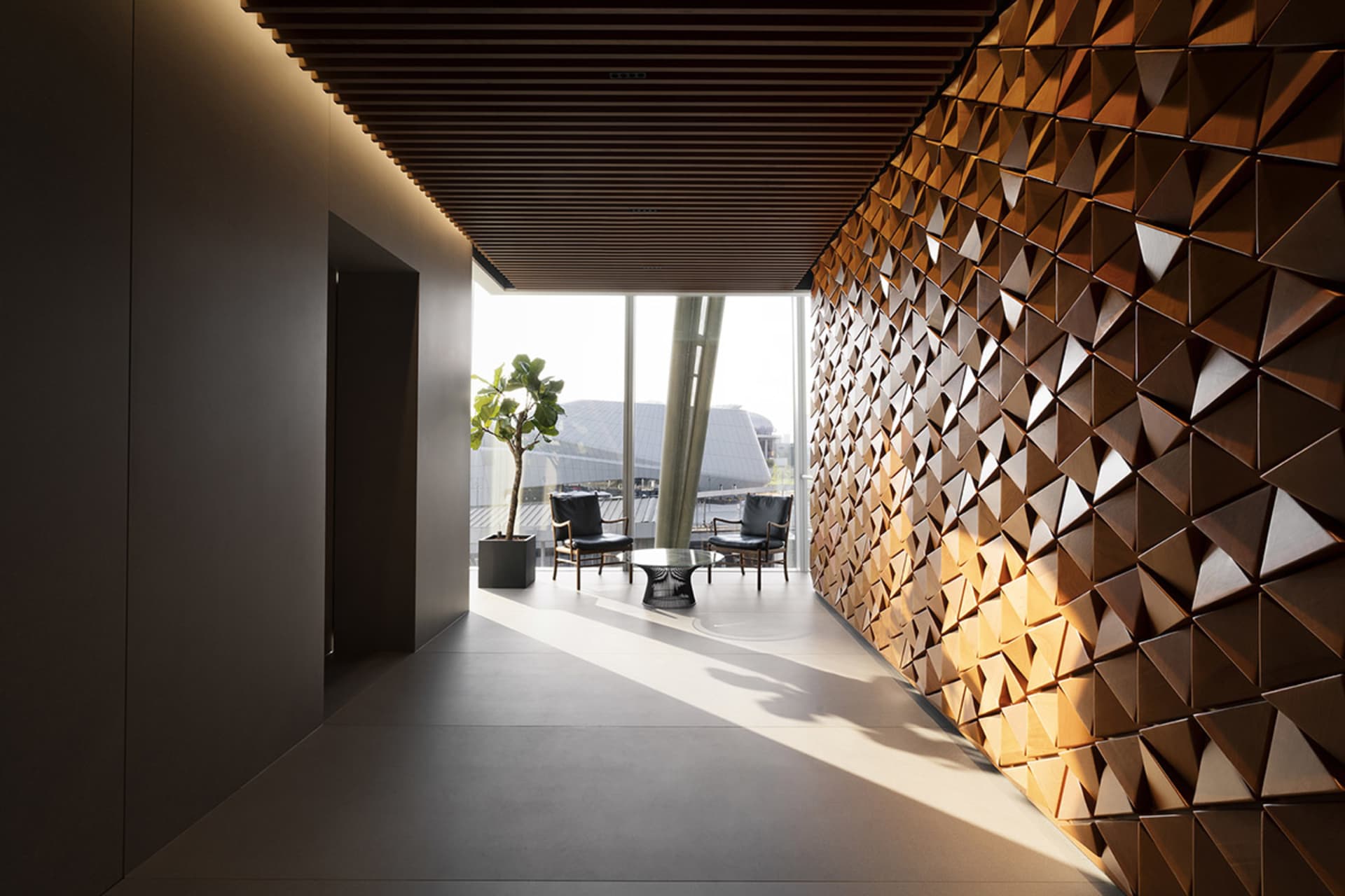 A modern office space featuring a wall made of wooden triangles and a small sitting area with chairs and a plant.