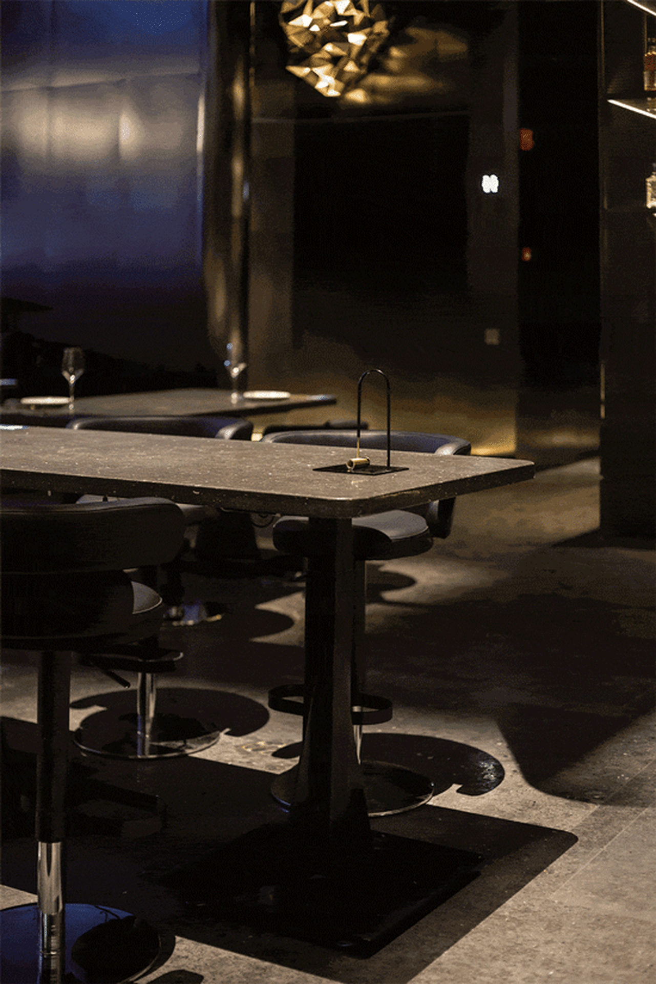 A modern restaurant bar with sleek, black metal tables and stools, illuminated by warm, ambient lighting.
