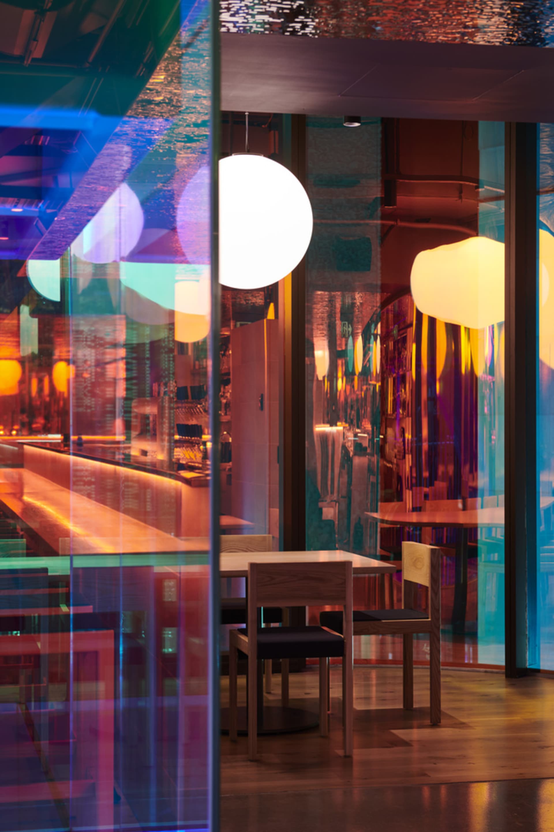 Modern restaurant interior with glass walls, illuminated tables and chairs, and colorful lighting effect.