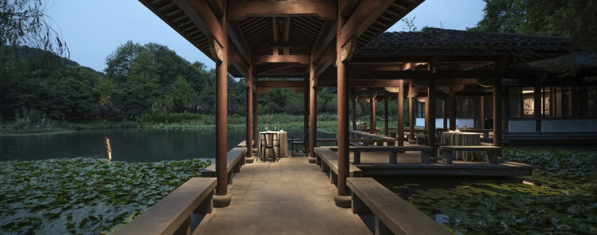 Outdoor seating area with wooden tables and chairs overlooking a serene lake surrounded by lush vegetation.