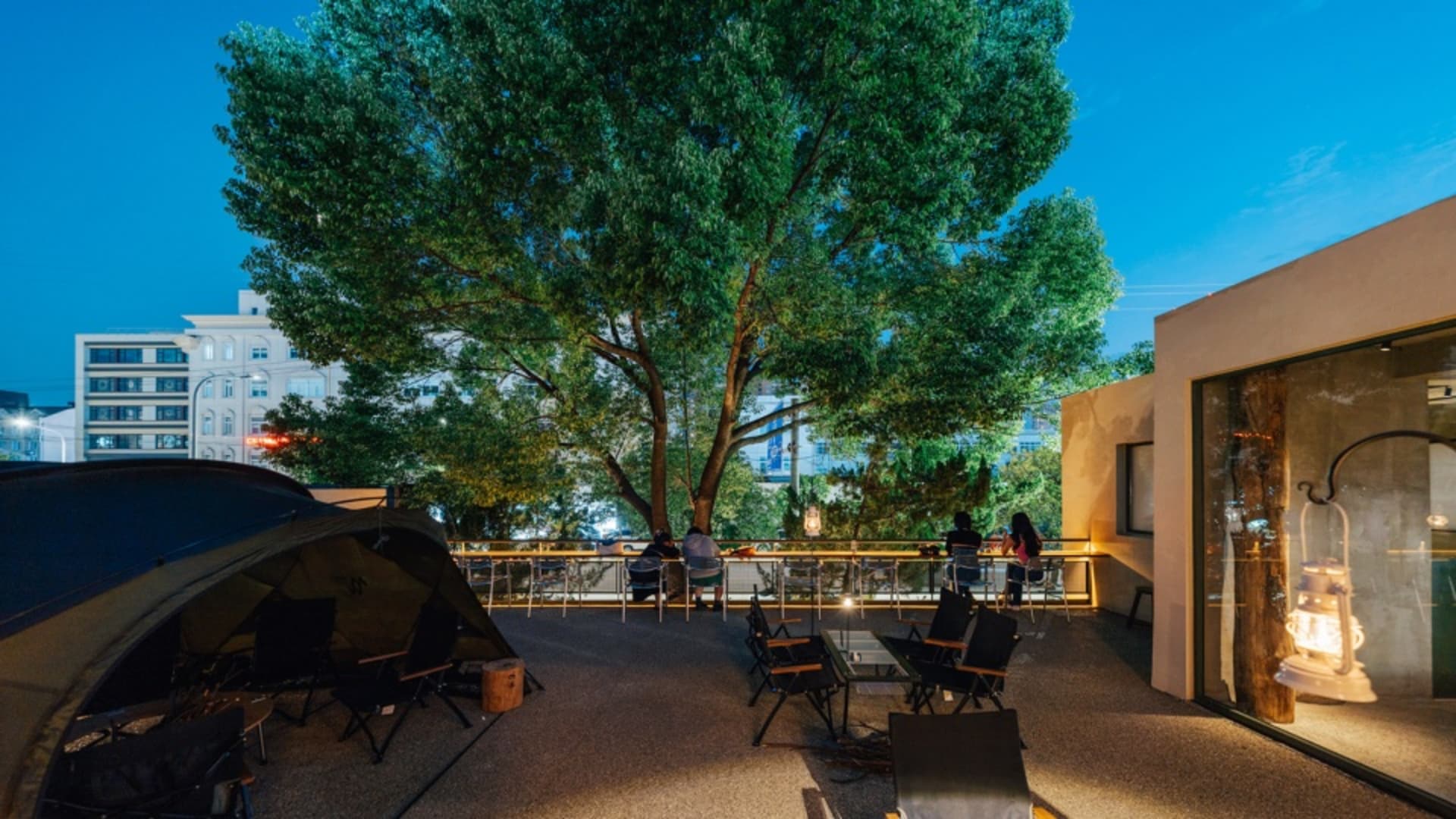 An outdoor seating area with a tent, chairs, and a lantern, surrounded by a tall tree and a modern building in the background