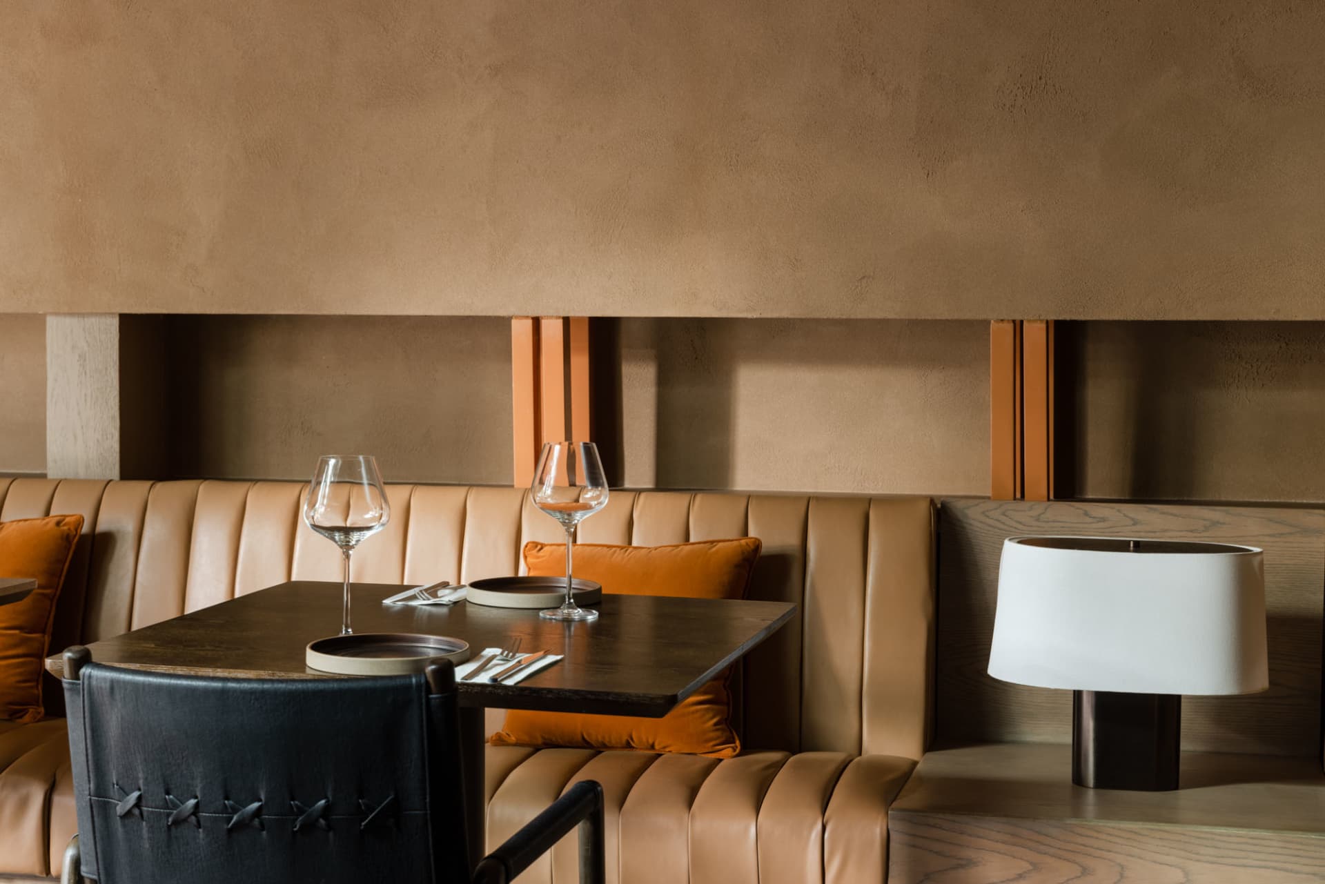 A modern restaurant booth featuring leather cushions and a wooden table with wine glasses, plates, and a lamp.