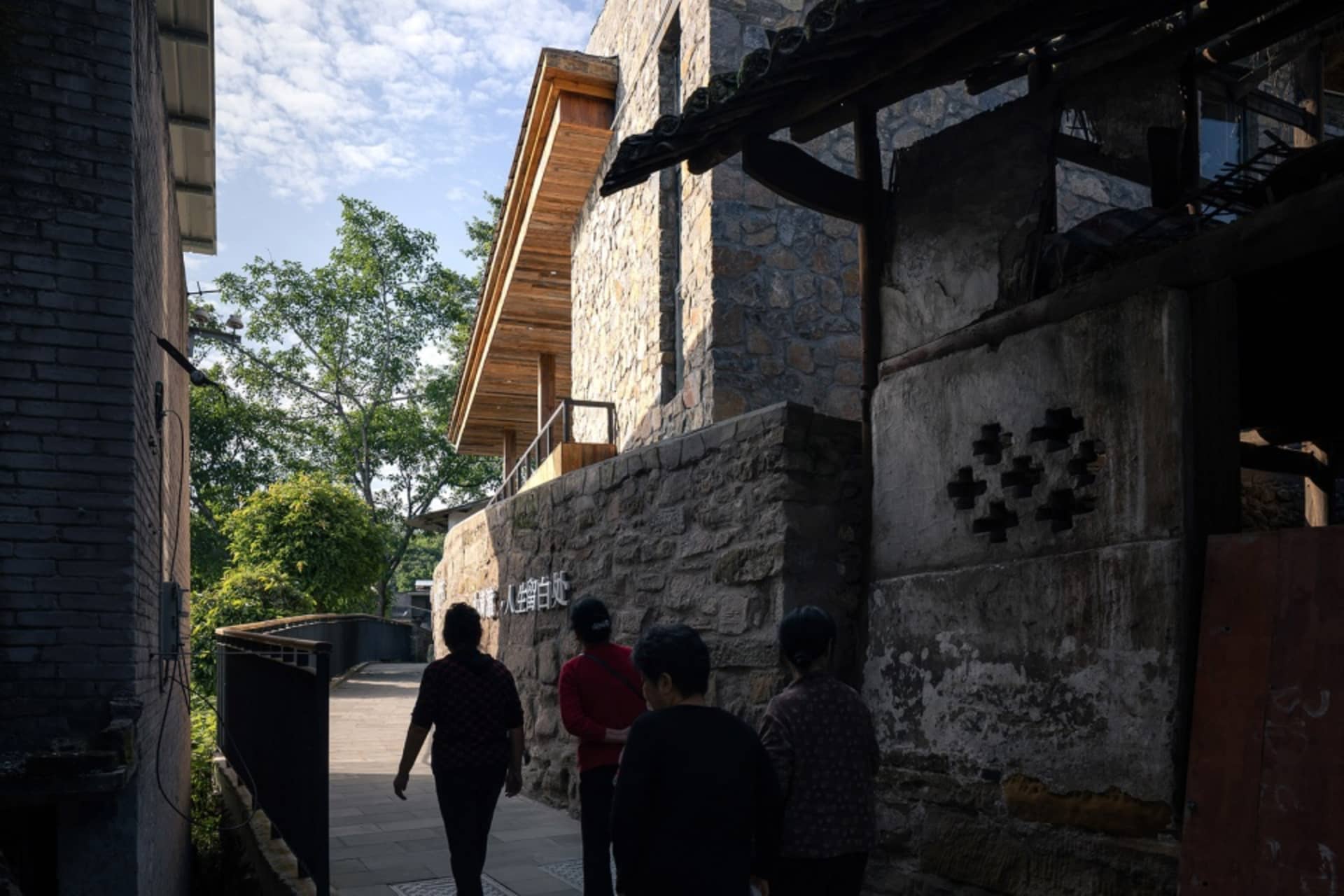 Three people walking down a quiet stone-walled street with trees and buildings.