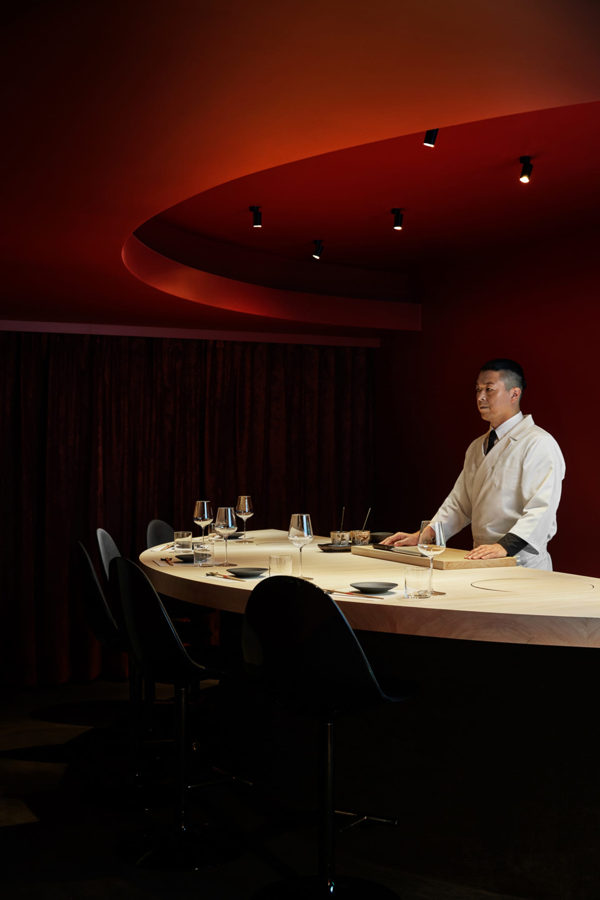 A chef stands at a modern restaurant counter with wood countertops and chairs.