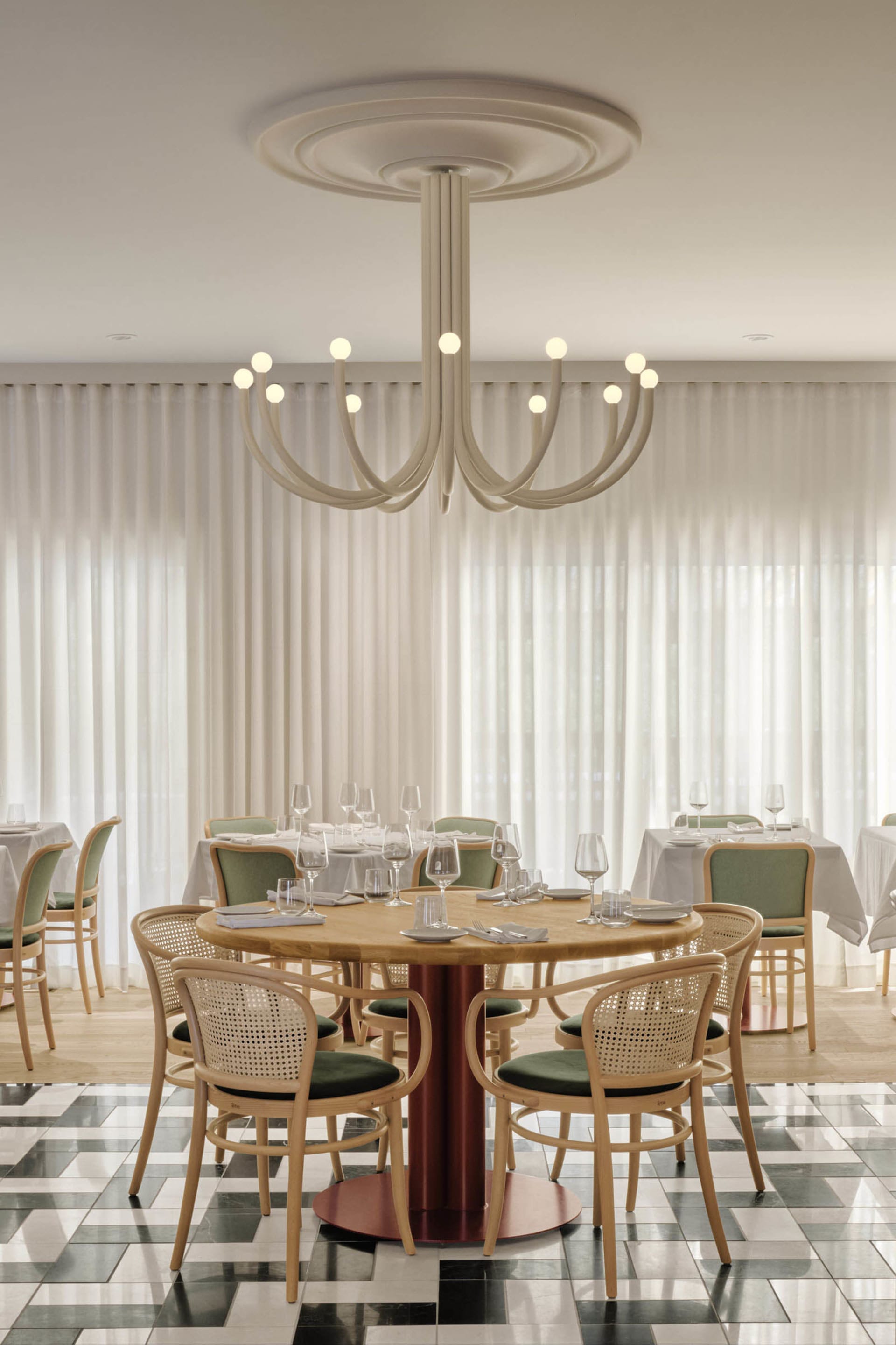 A modern dining room with wooden tables and chairs, white curtains, and a white chandelier.