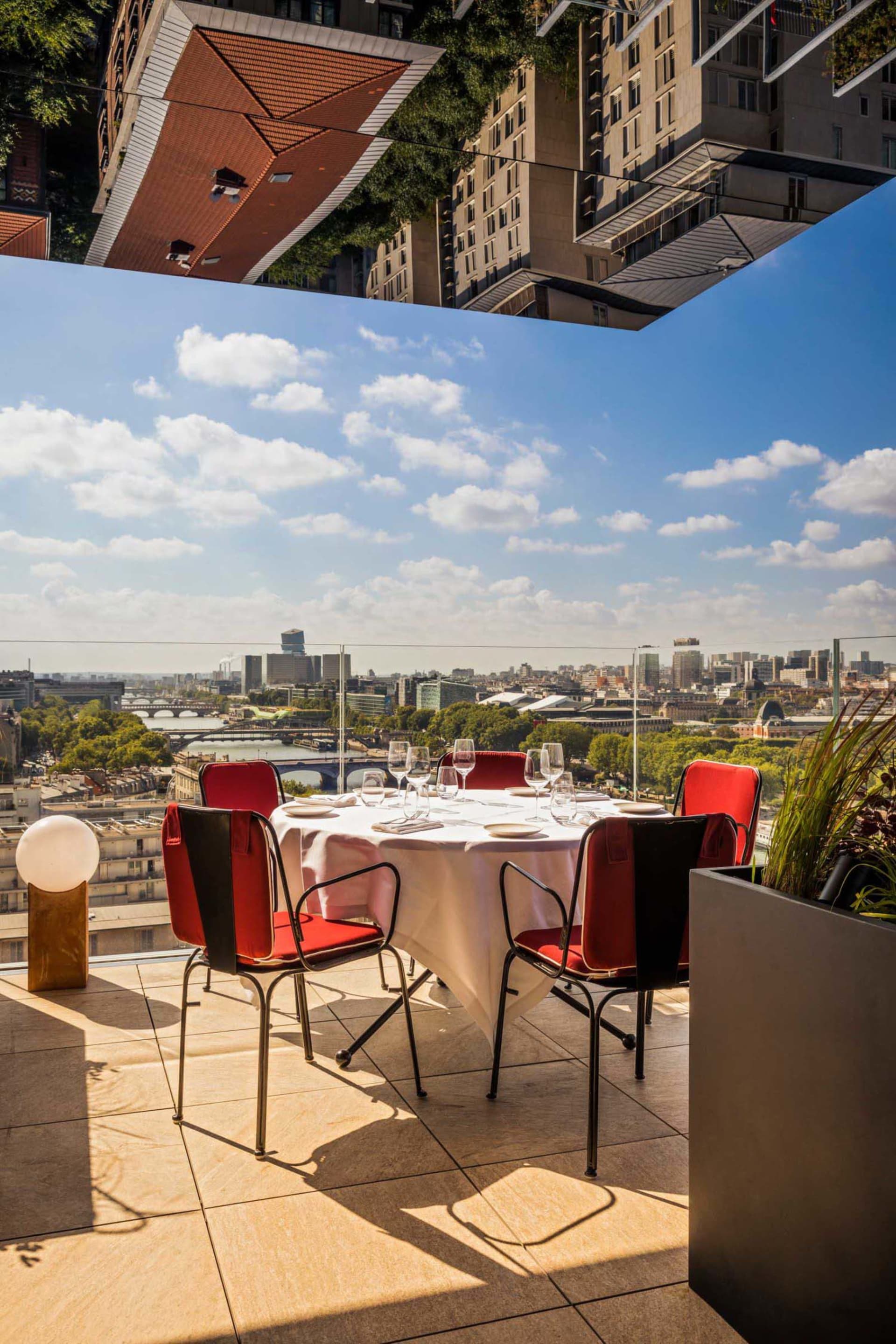 A rooftop dining table set with wine glasses and plates, overlooking a cityscape under a clear sky.