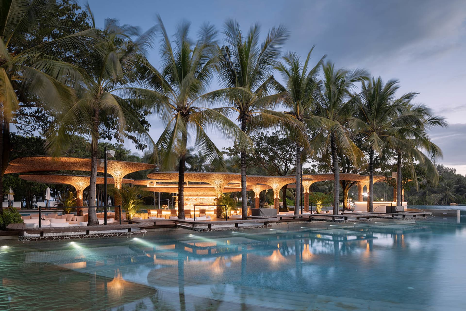 A beautiful tropical poolside bar illuminated with warm lighting, surrounded by palm trees at dusk.