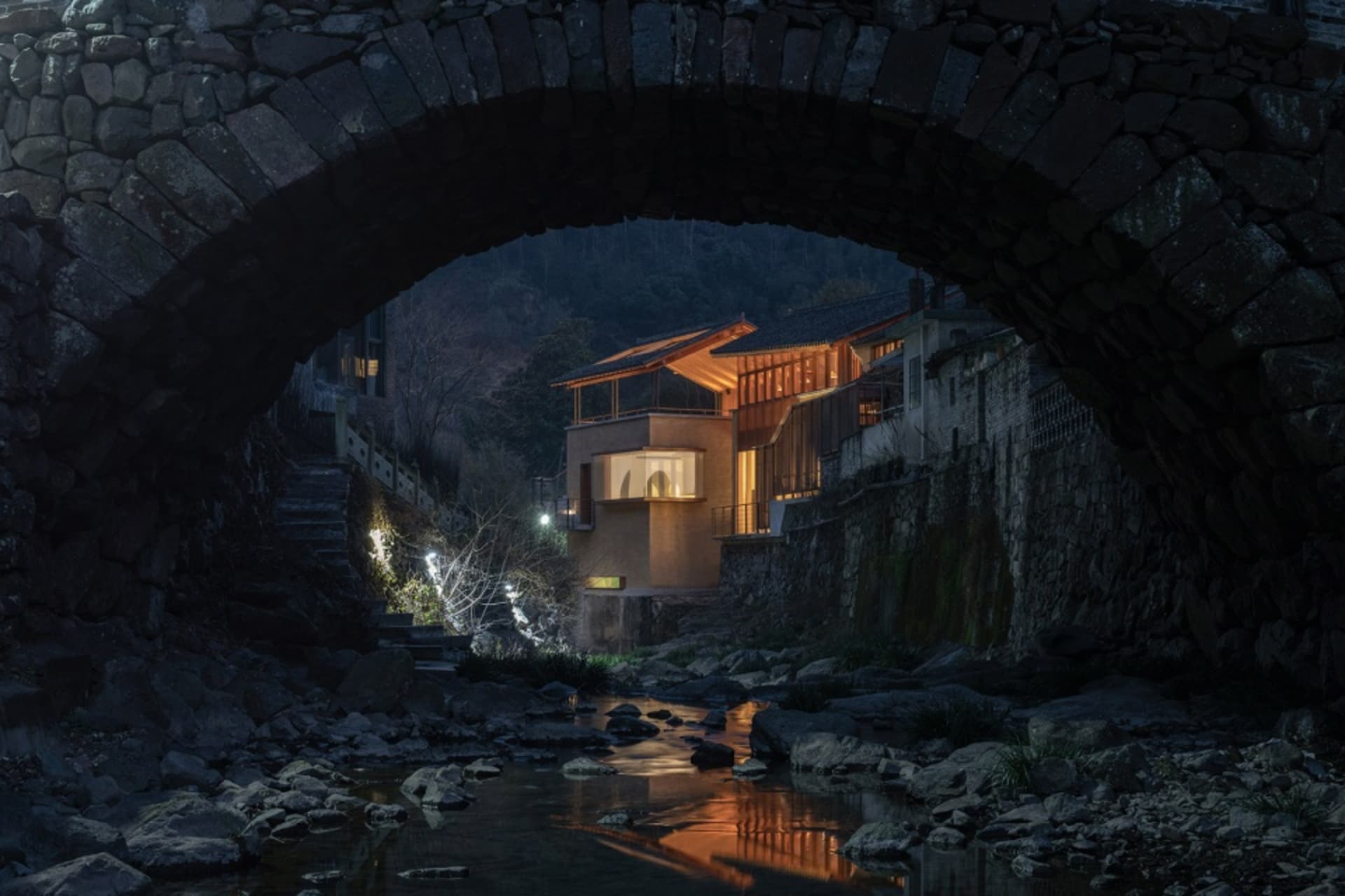 夜景中的石材與混凝土建築住宅，搭配木質元素，並以溫暖燈光照亮。