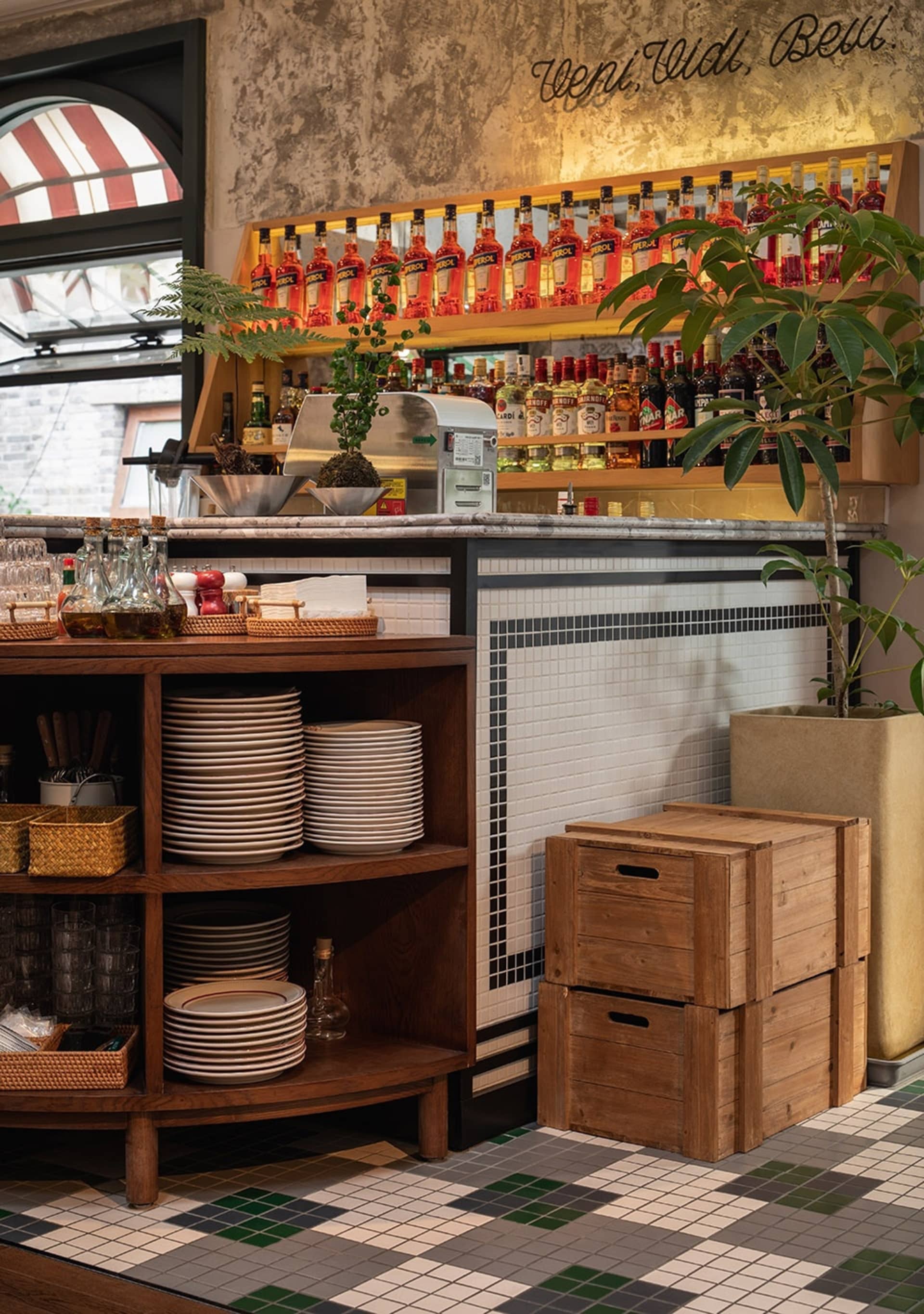 A coffee shop's retail counter featuring a wooden shelf with plates, metal bar counter, and potted plants.