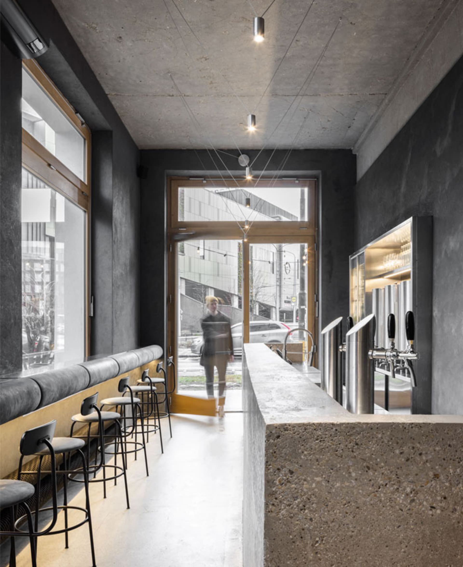 A modern, industrial-style bar with a concrete counter and seating area, featuring a minimalist design and a glass door.